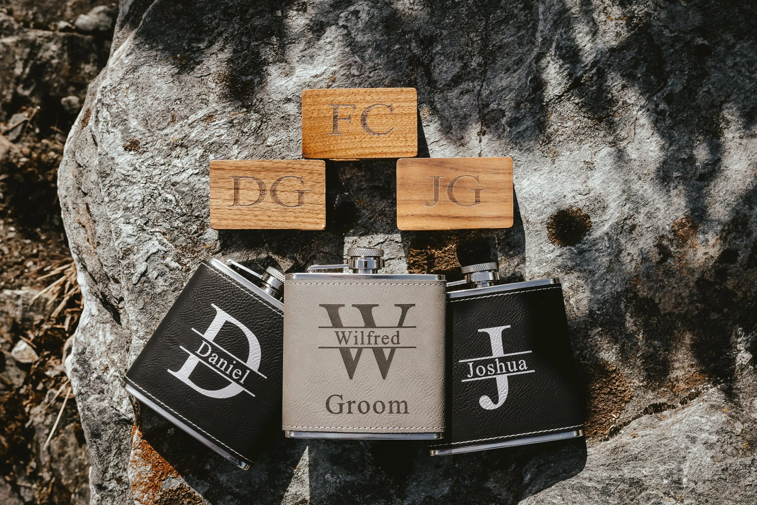 Three wooden tiles with engraved initials 'FC', 'DG', and 'JG' placed on a rock, with two small lenses and a '_W' groom badge nearby.