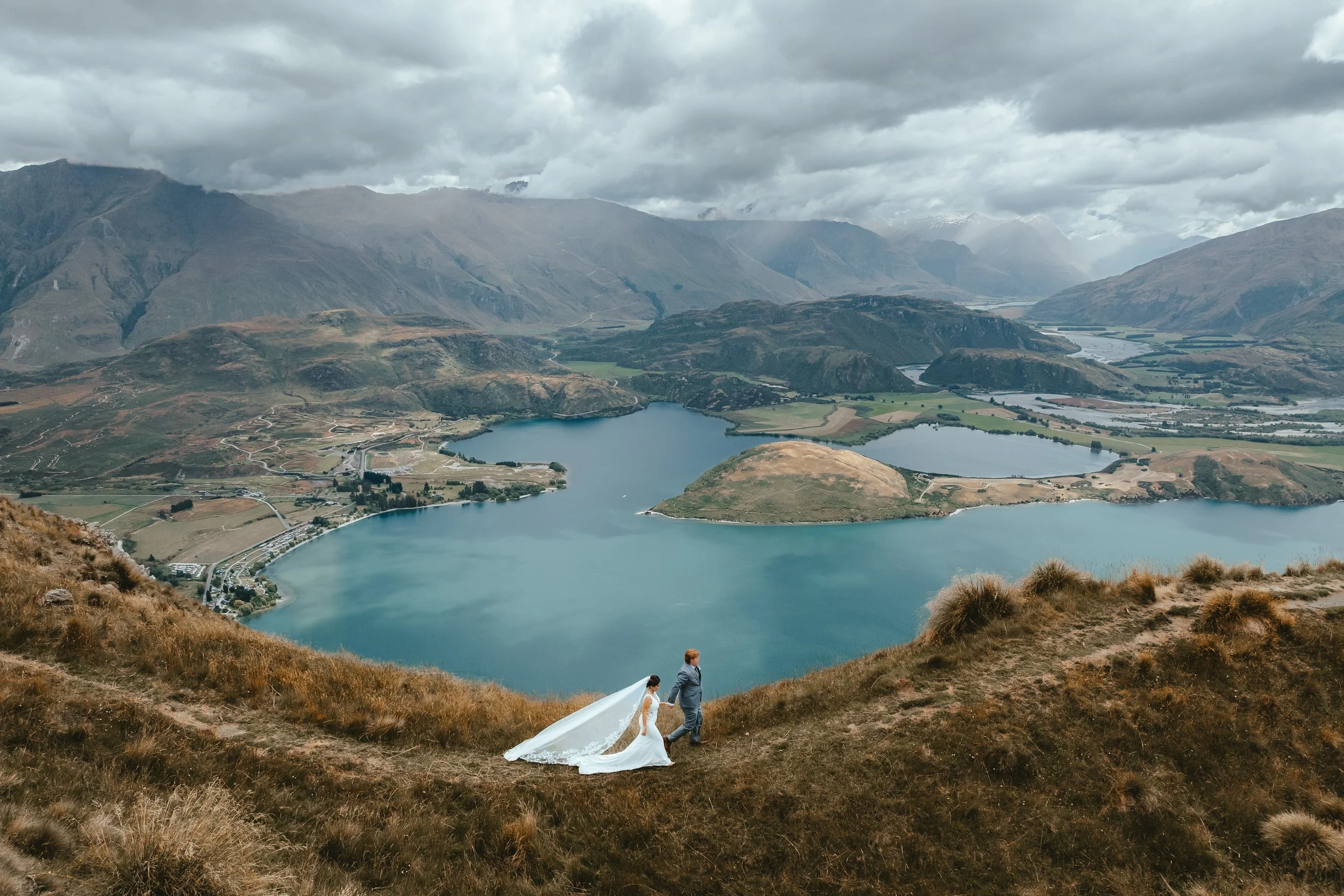 A bride and groom walking down a grassy hillside overlooking a lake and mountains, with a cloudy sky overhead.