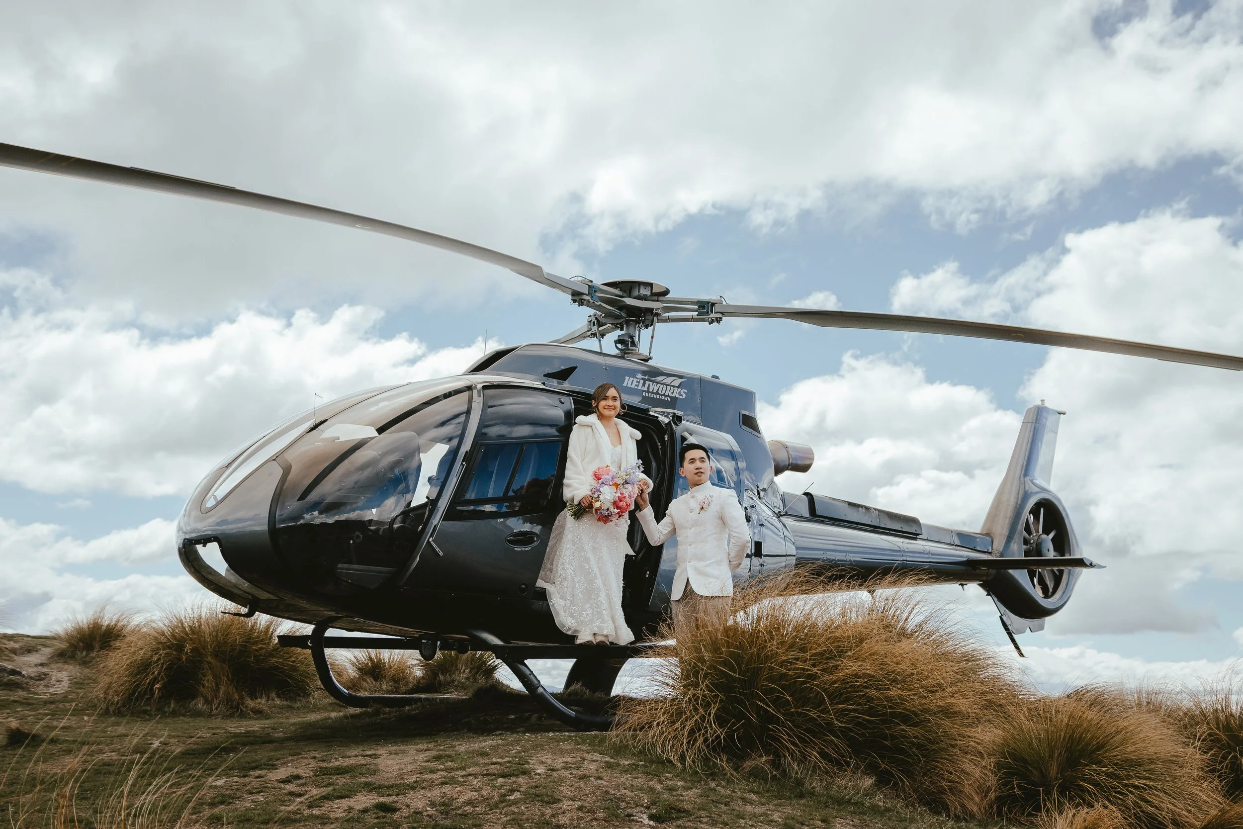 A bride and groom standing next to a black helicopter decorated for a wedding, outdoors on grassy terrain with cloudy sky in the background.