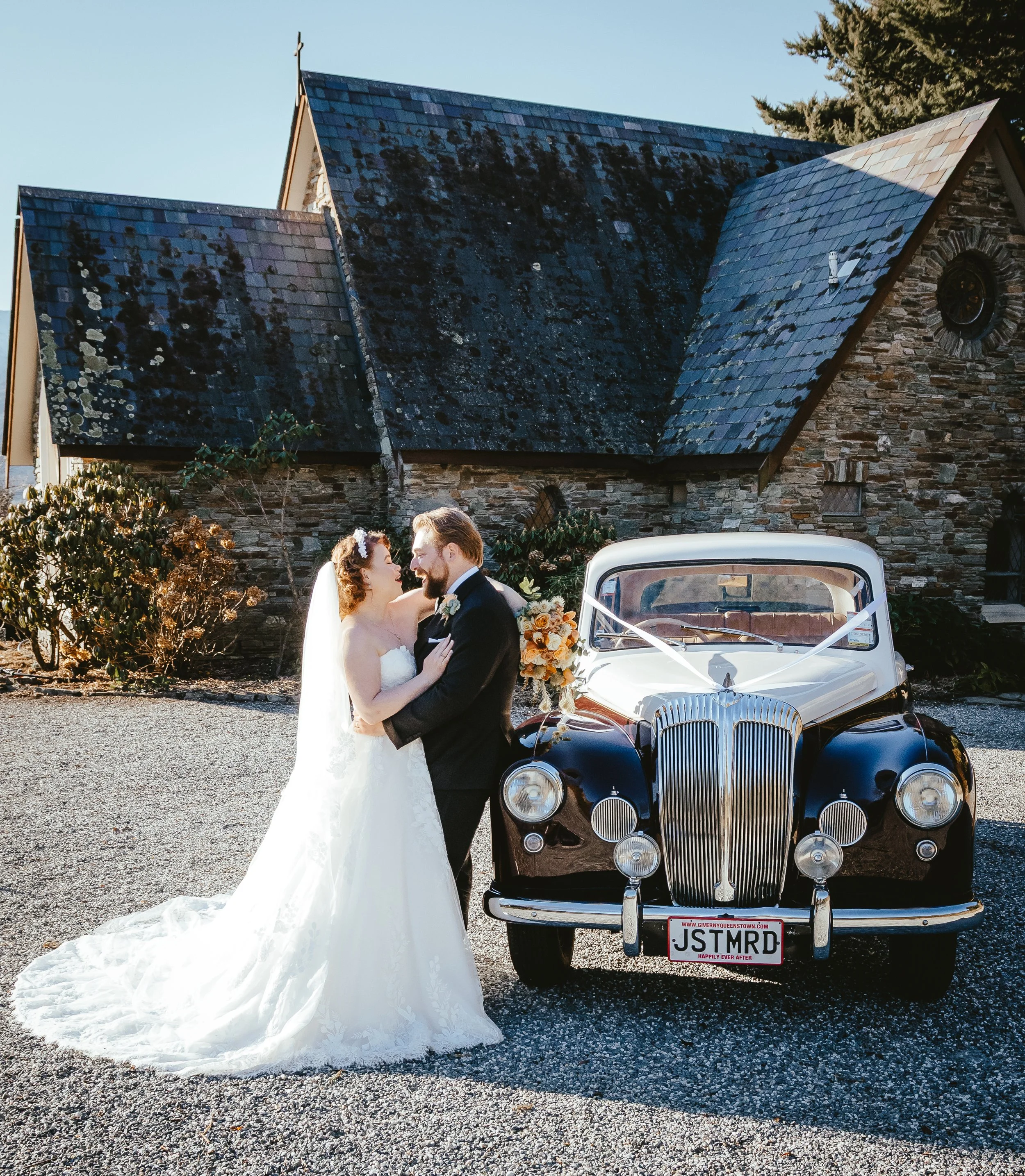 A bride and groom stand close together in front of a vintage black car decorated for a wedding, with a church building in the background. The bride wears a white wedding dress and veil, and the groom is in a black suit. They are smiling and looking a