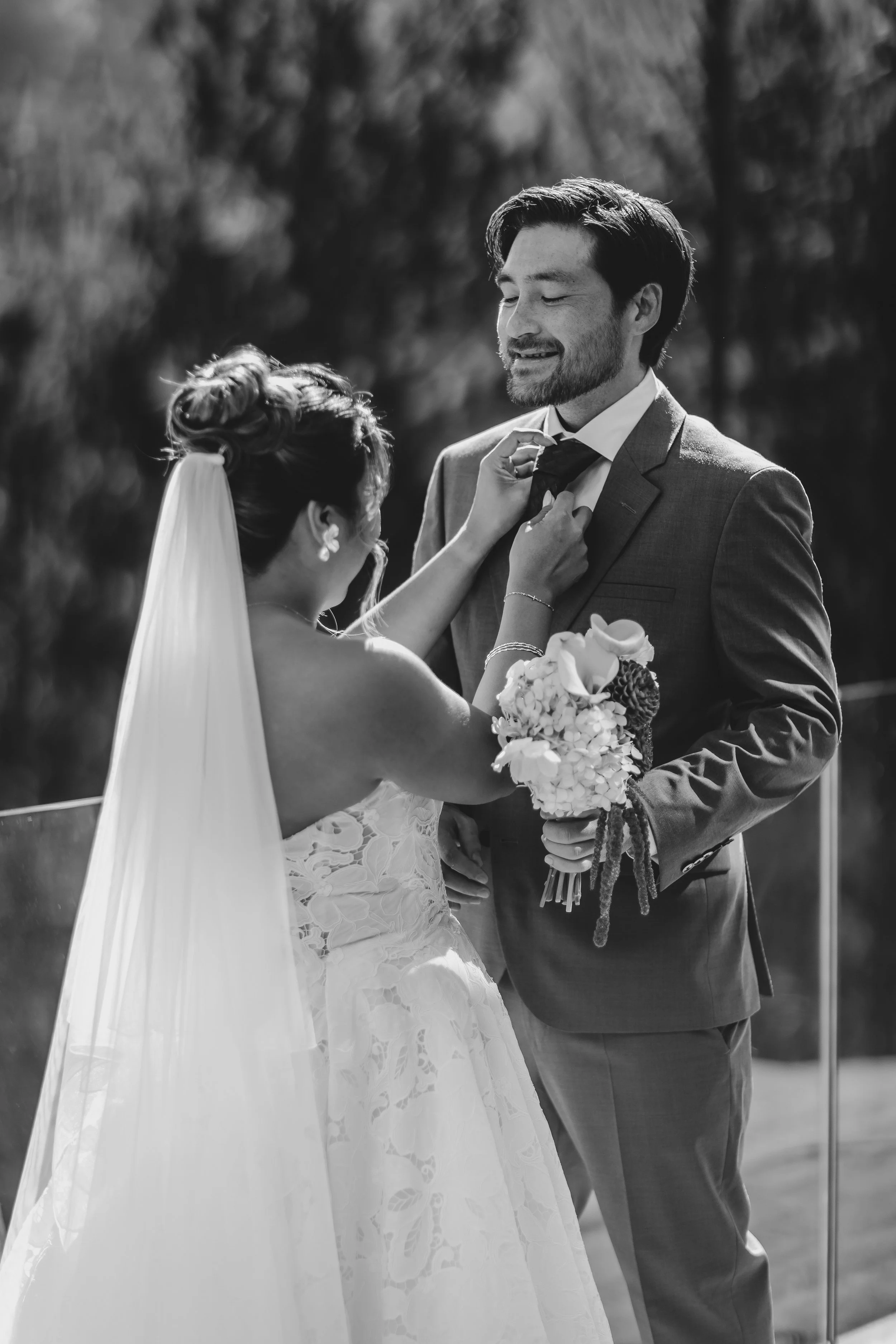 A bride and groom at their wedding, with the bride adjusting the groom's tie and holding a bouquet of flowers.
