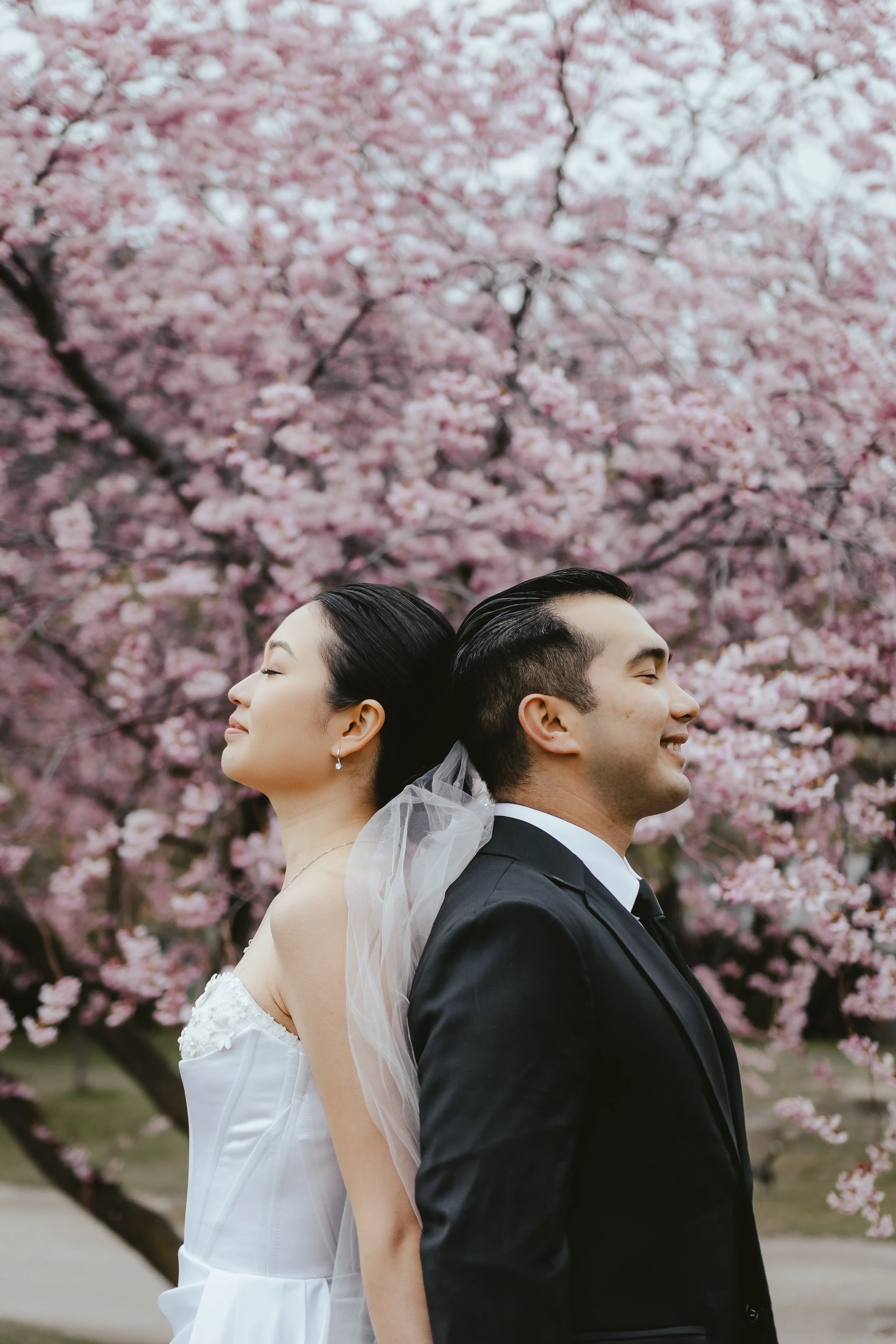 A bride and groom are standing back to back outdoors, eyes closed, smiling softly, with a background of pink cherry blossom trees.