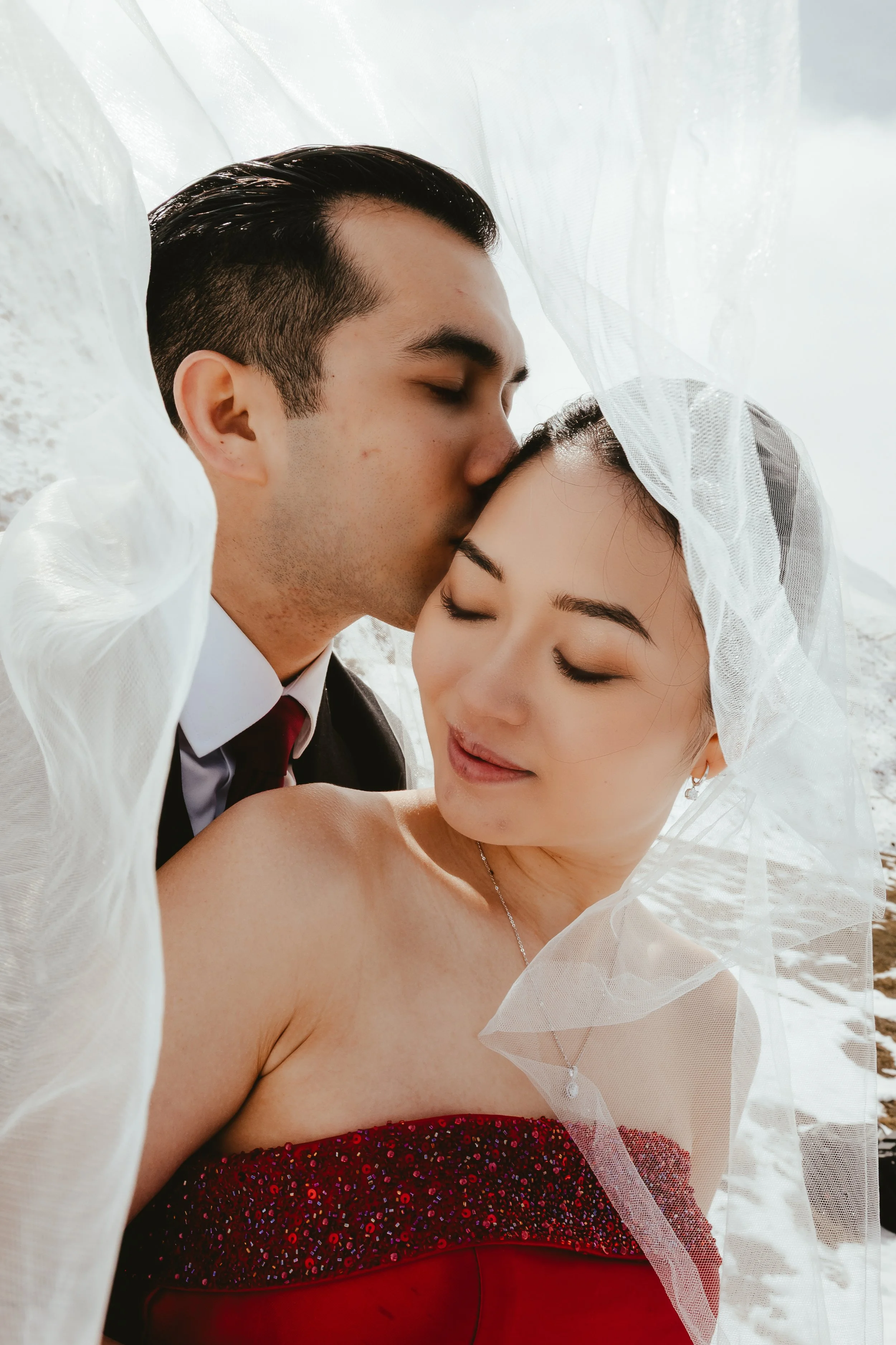 A close-up photo of a couple with eyes closed, sharing a tender moment, with the man kissing the woman's forehead. The man has short, dark, styled hair and is wearing a tuxedo with a white shirt and dark tie, while the woman has dark hair and is wear