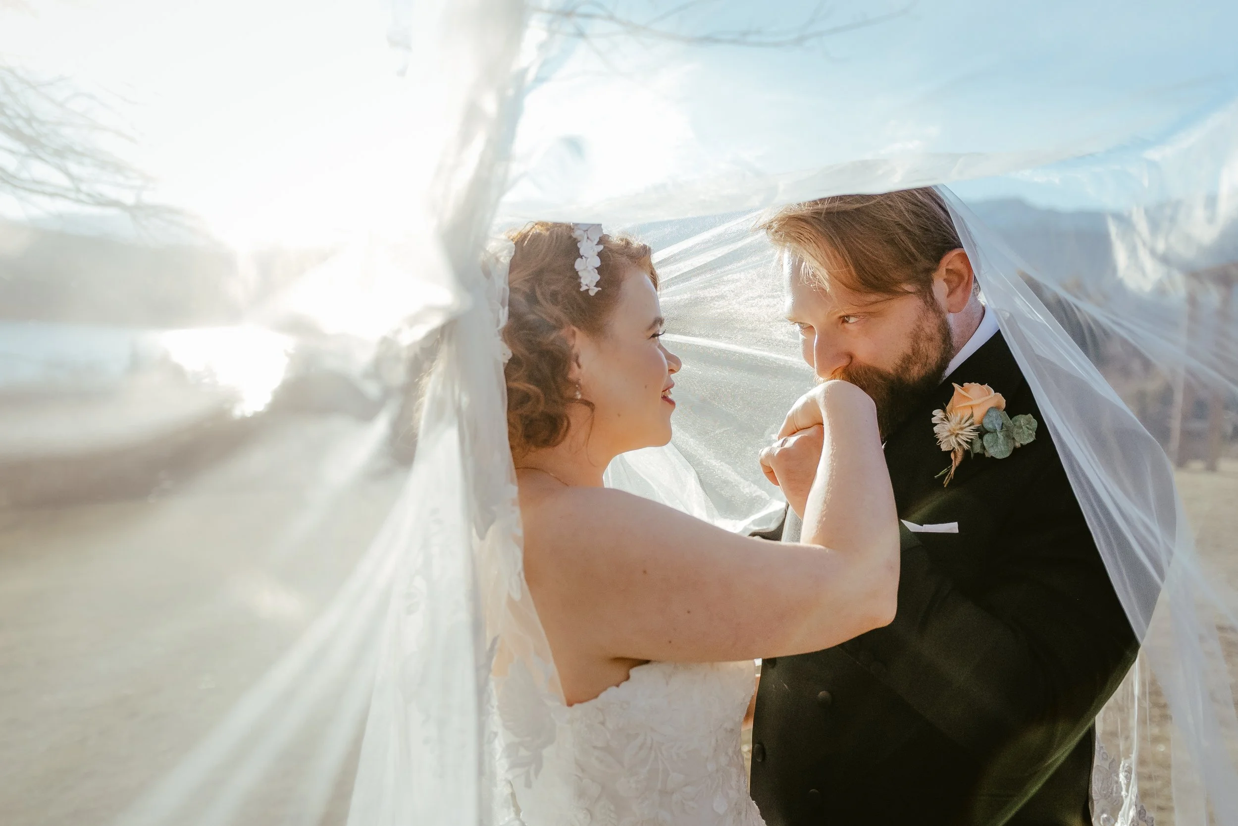 A bride and groom under a veil, looking into each other's eyes during their wedding ceremony on a sunny outdoor day.