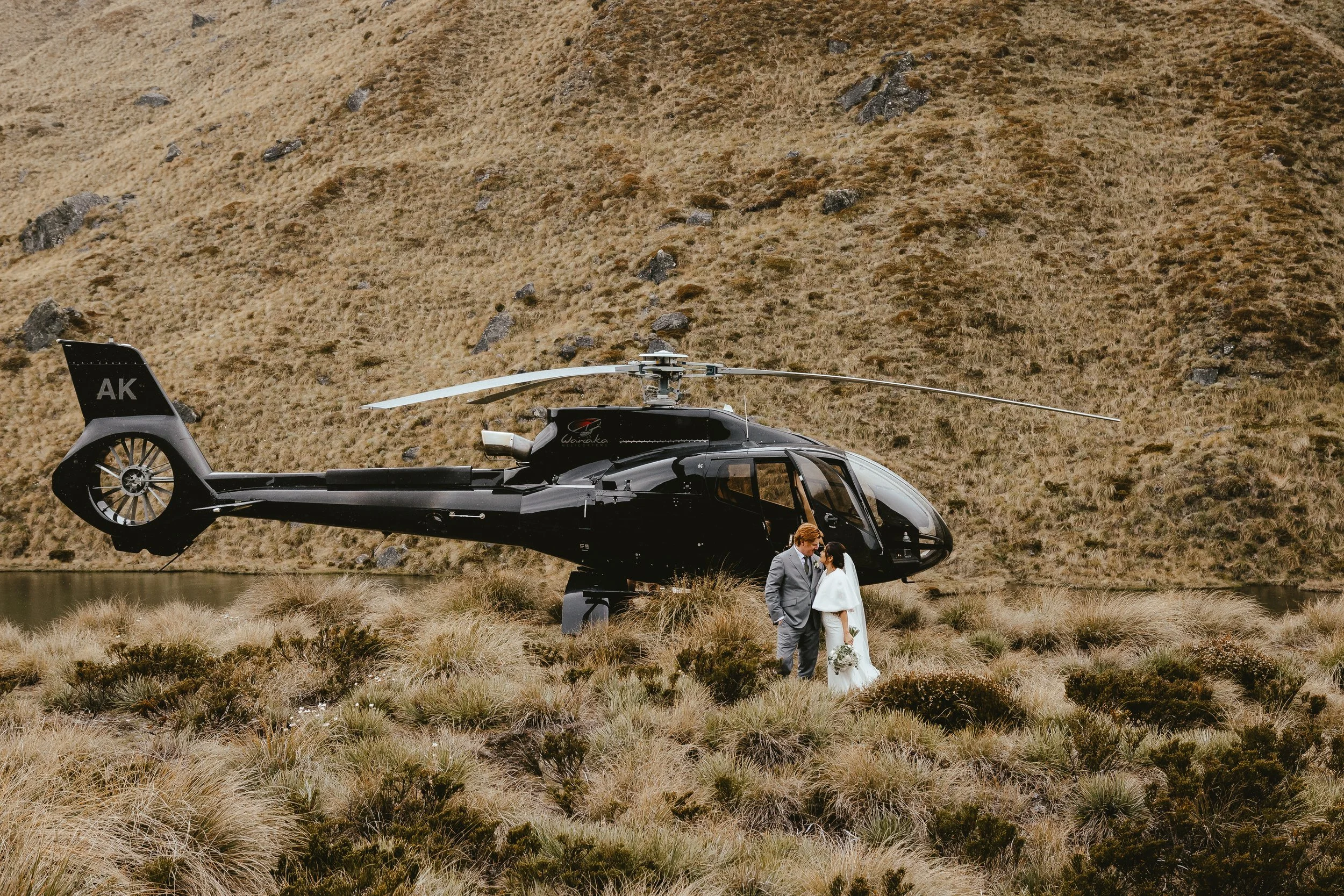 A couple in wedding attire standing in front of a black helicopter in a grassy, hilly landscape.