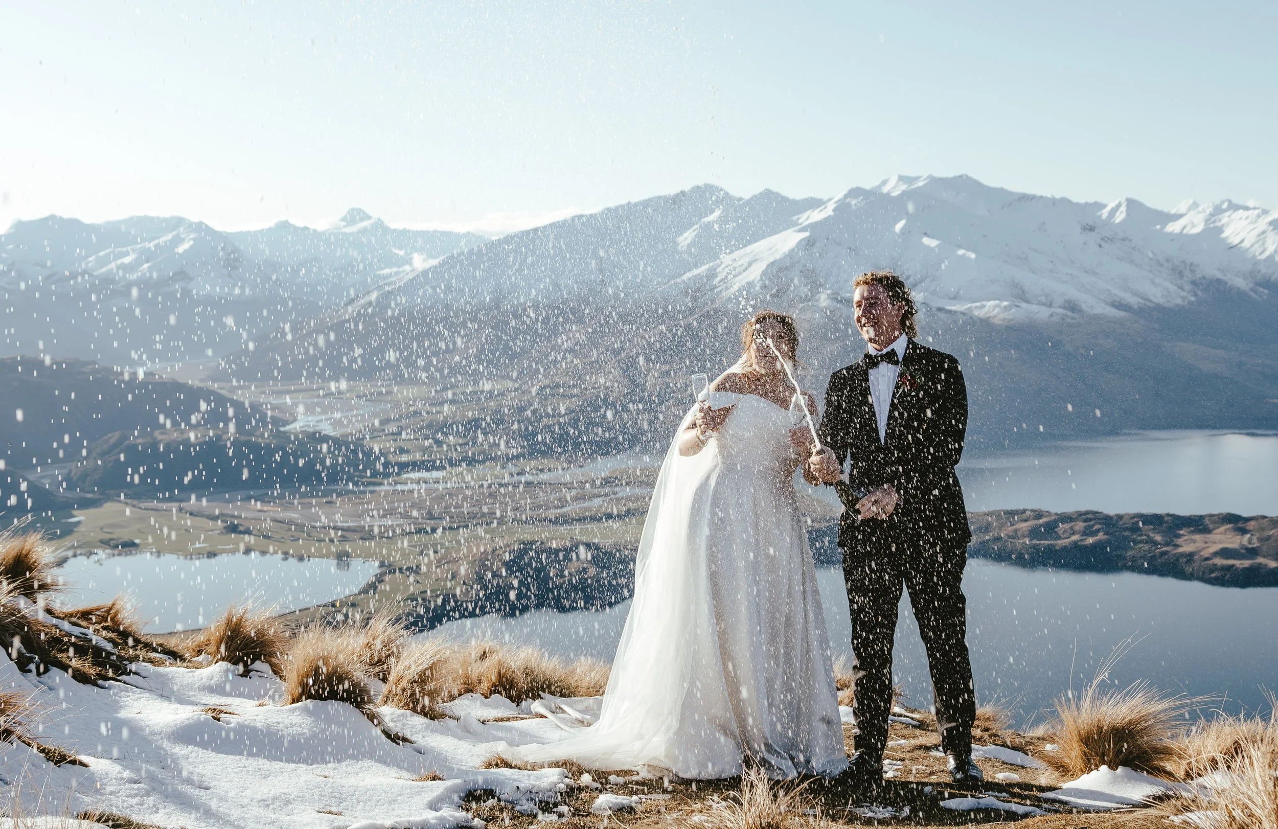 A bride and groom in formal attire celebrate outdoors near snow-capped mountains, spraying champagne in a joyful toast.