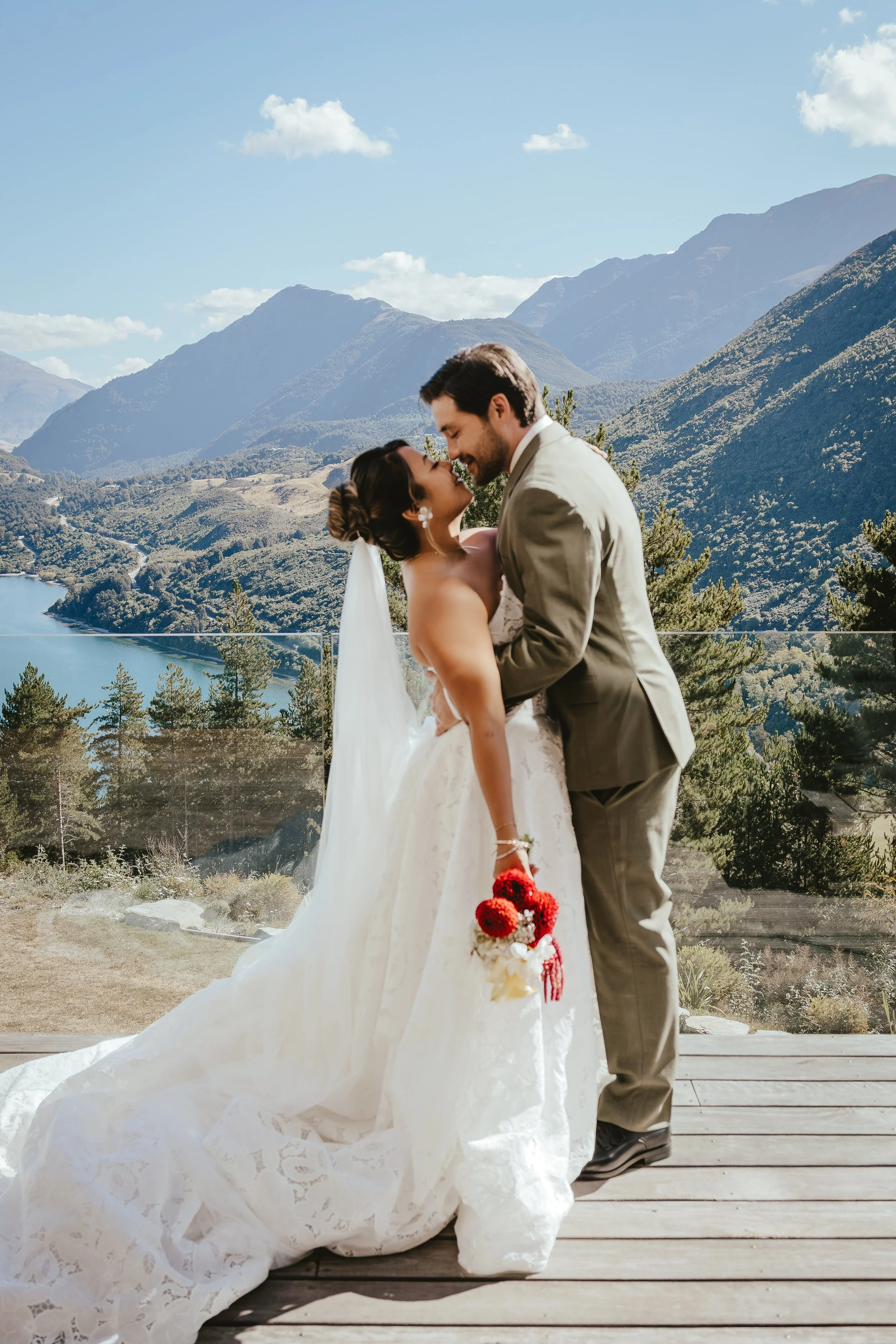 A bride and groom sharing a romantic moment outdoors on a wooden deck with scenic mountains and a lake in the background. The bride is holding a bouquet of red and cream flowers, and both are smiling with their foreheads touching.