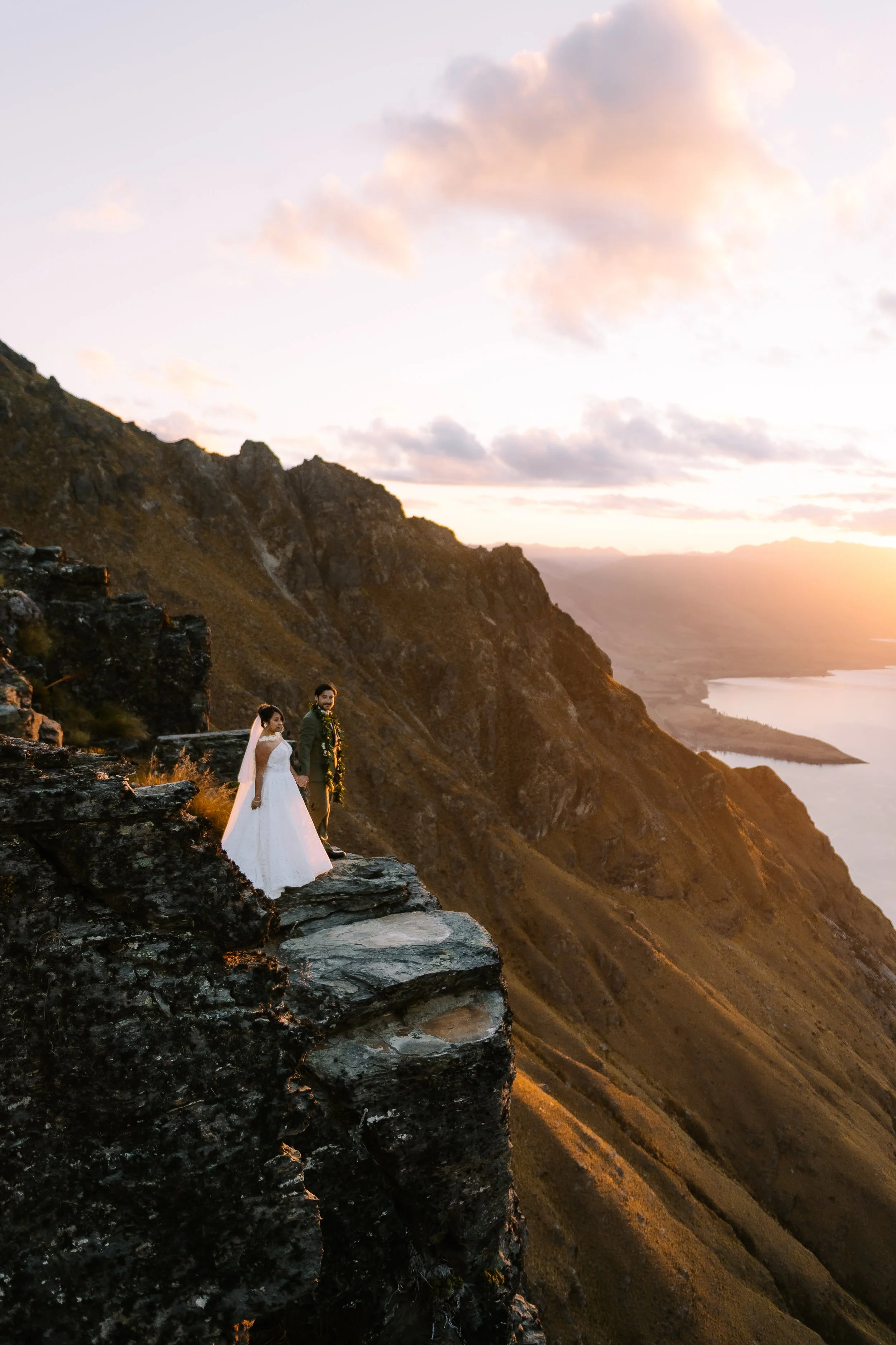 A bride and groom standing on a rocky ledge overlooking a mountain and river landscape at sunset.