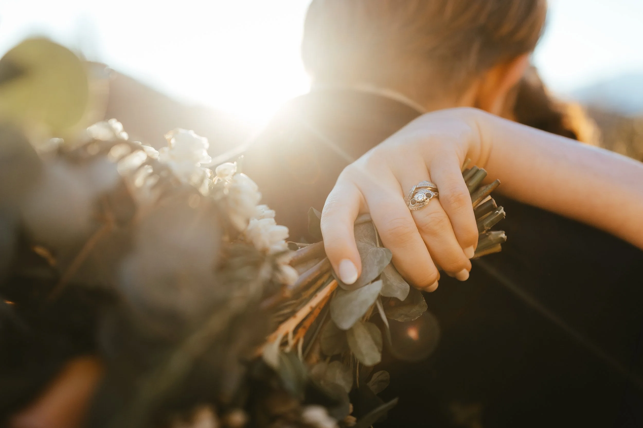 Close-up of a person's hand with a silver ring, holding a bundle of sticks or herbs, with sunlight in the background.