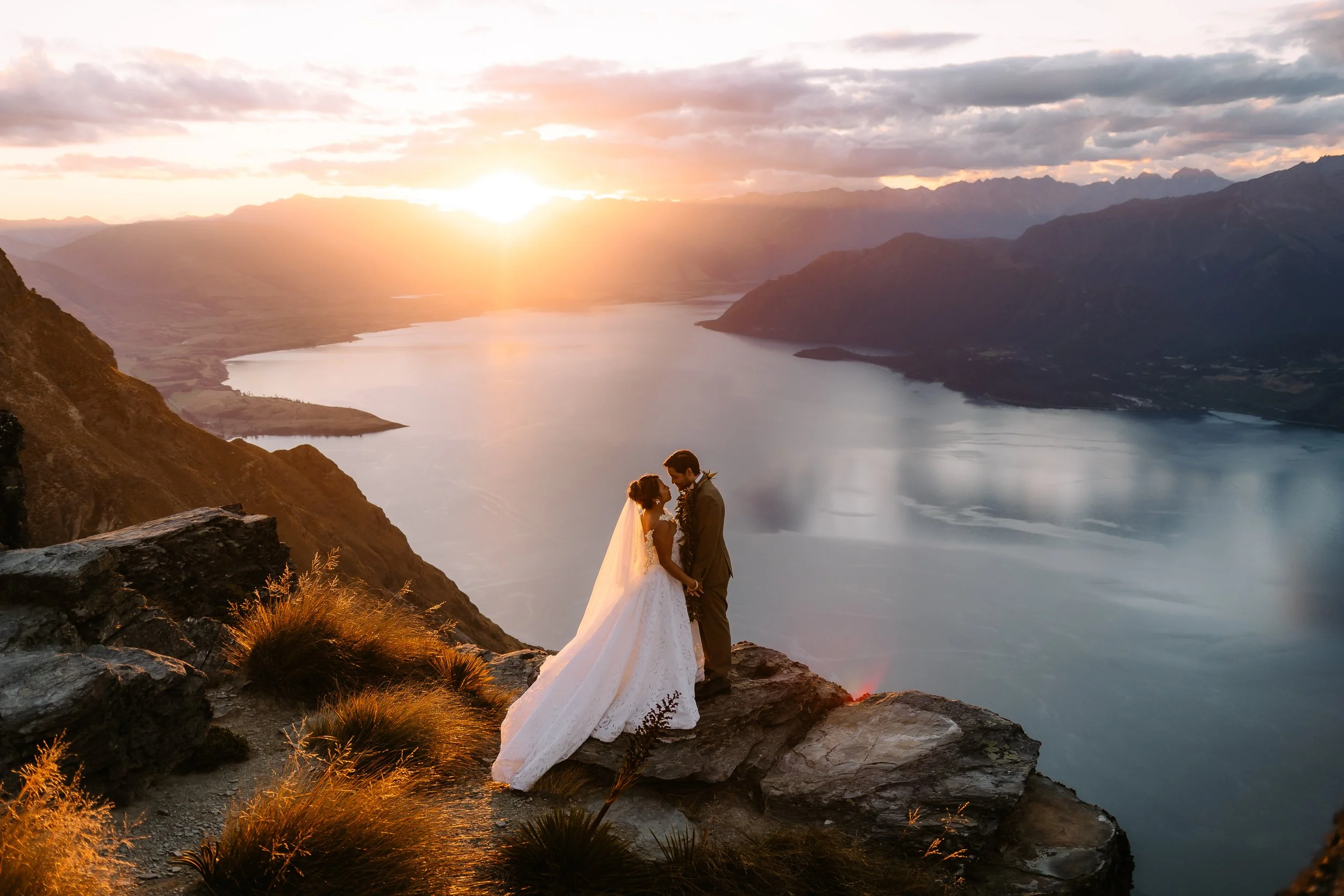 A bride and groom holding hands and looking at each other on a rocky ledge overlooking a lake and mountain range at sunset.