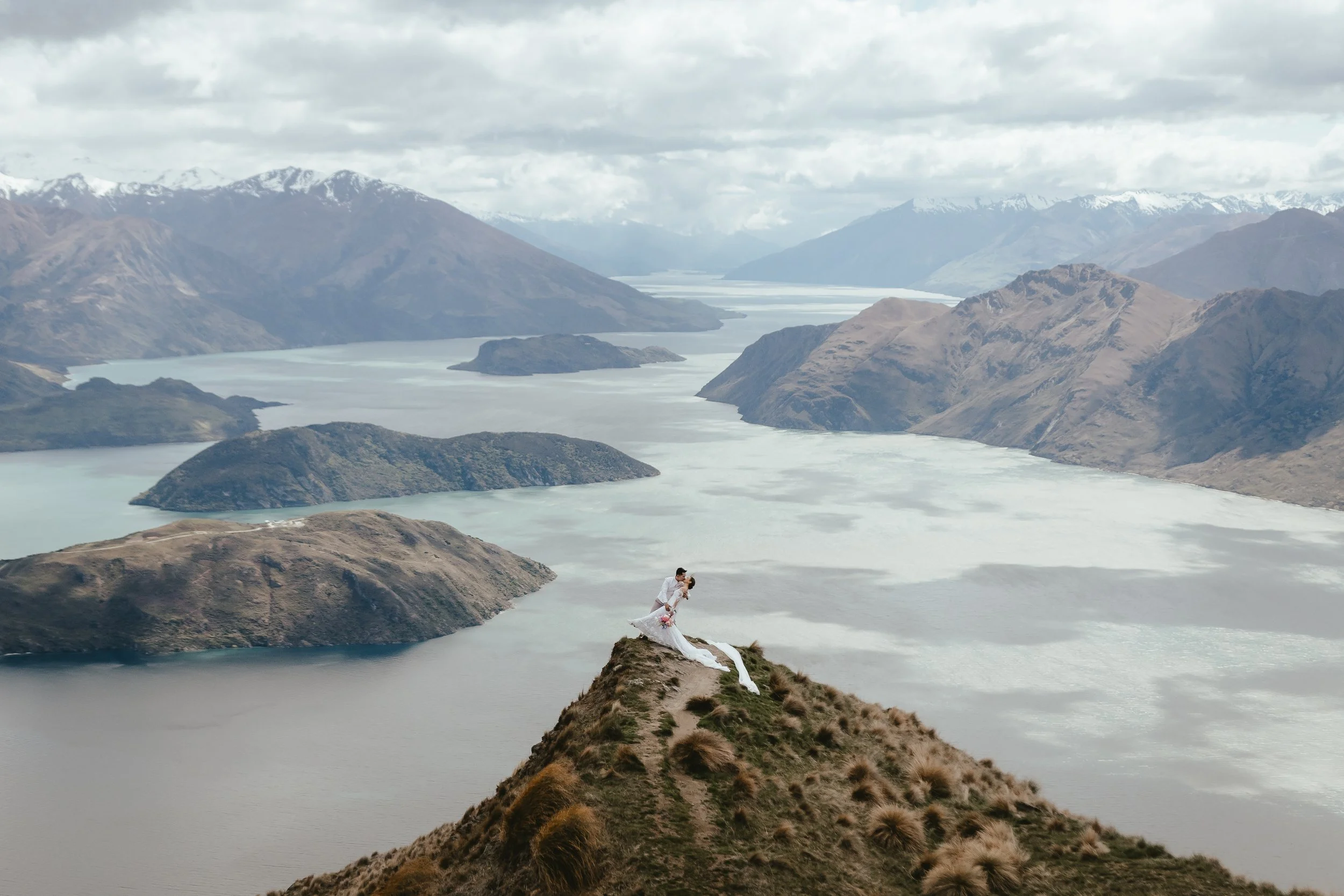 A bride and groom stand on a hill overlooking a large lake with mountains in the background, during overcast weather.