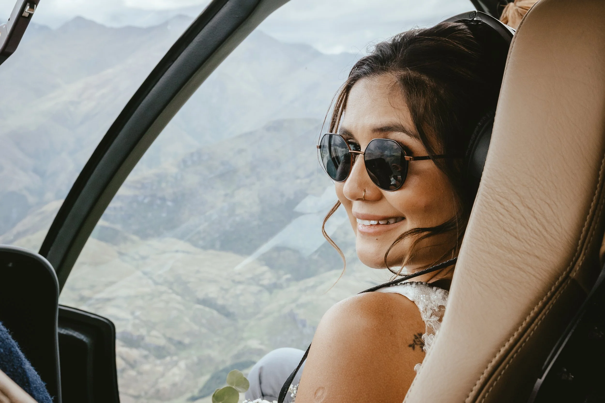 A woman with dark hair, wearing sunglasses and headphones, smiling while sitting in an aircraft with window view of mountains.
