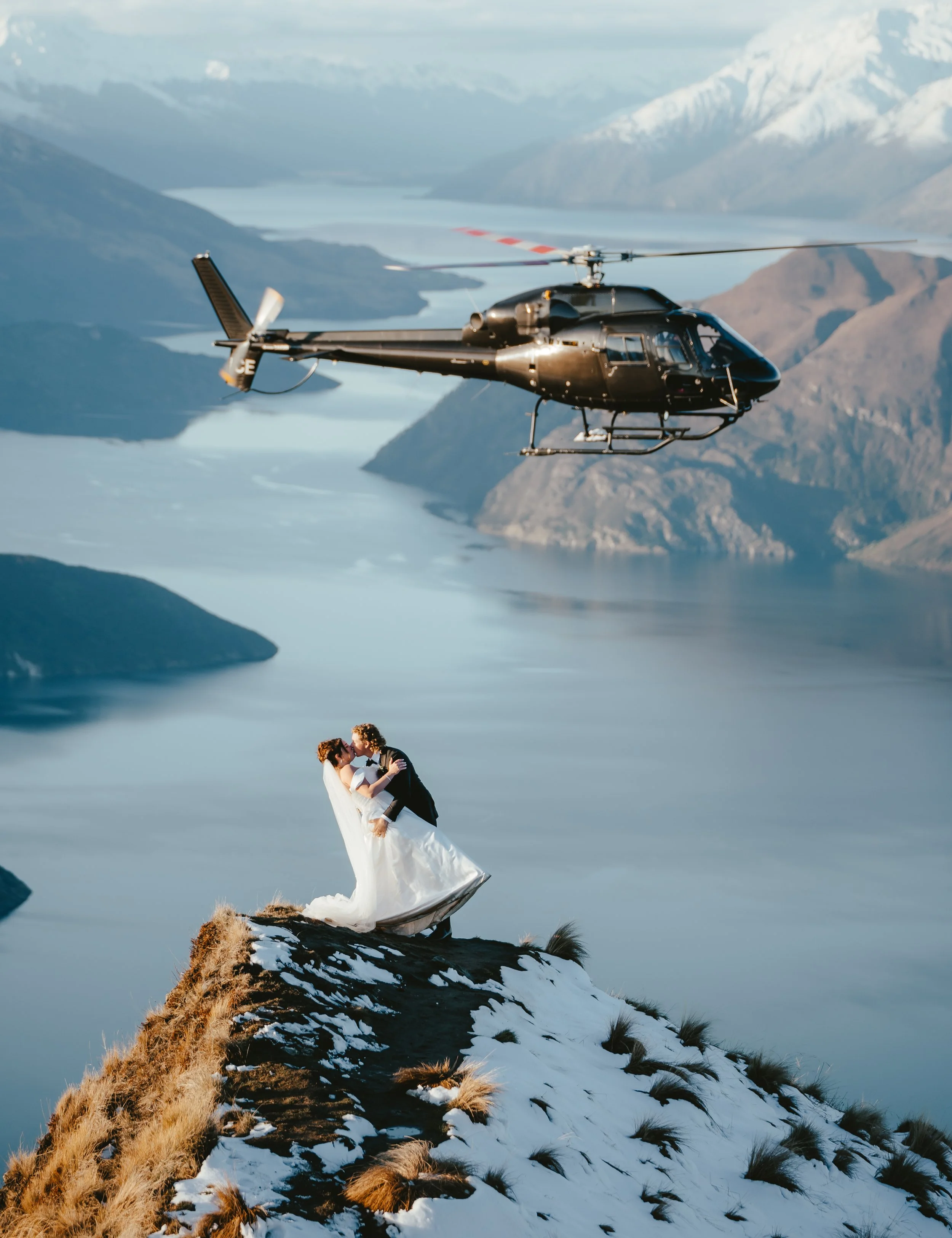A helicopter flying over a snowy mountain with a couple in wedding attire standing on a rocky ledge.