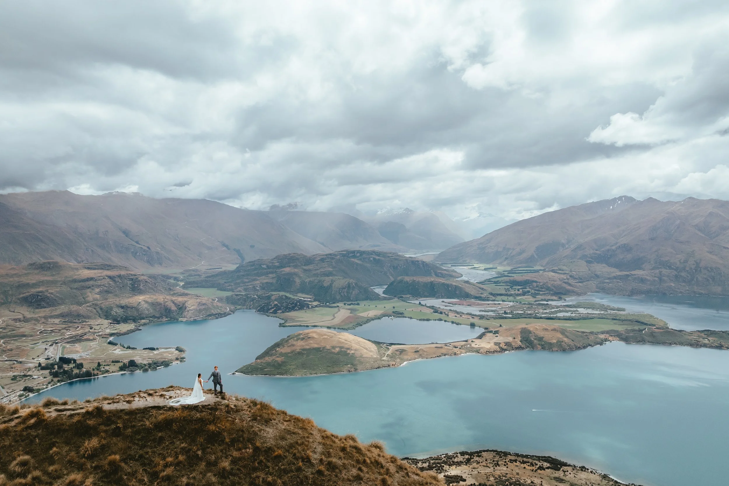 A bride and groom standing hand in hand on a hill overlooking a large lake and mountainous landscape under cloudy skies.