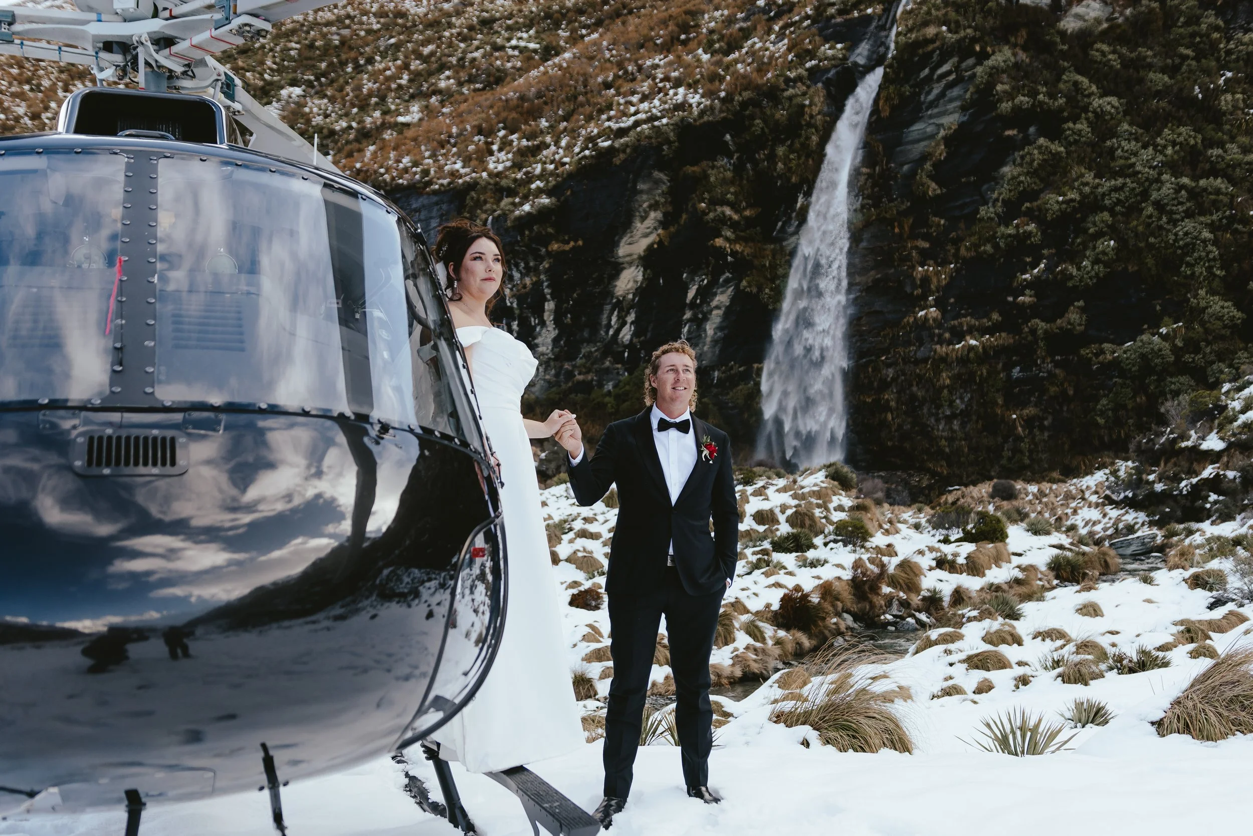 A bride and groom standing outdoors in snowy terrain beside a helicopter with mountain scenery and a waterfall in the background.