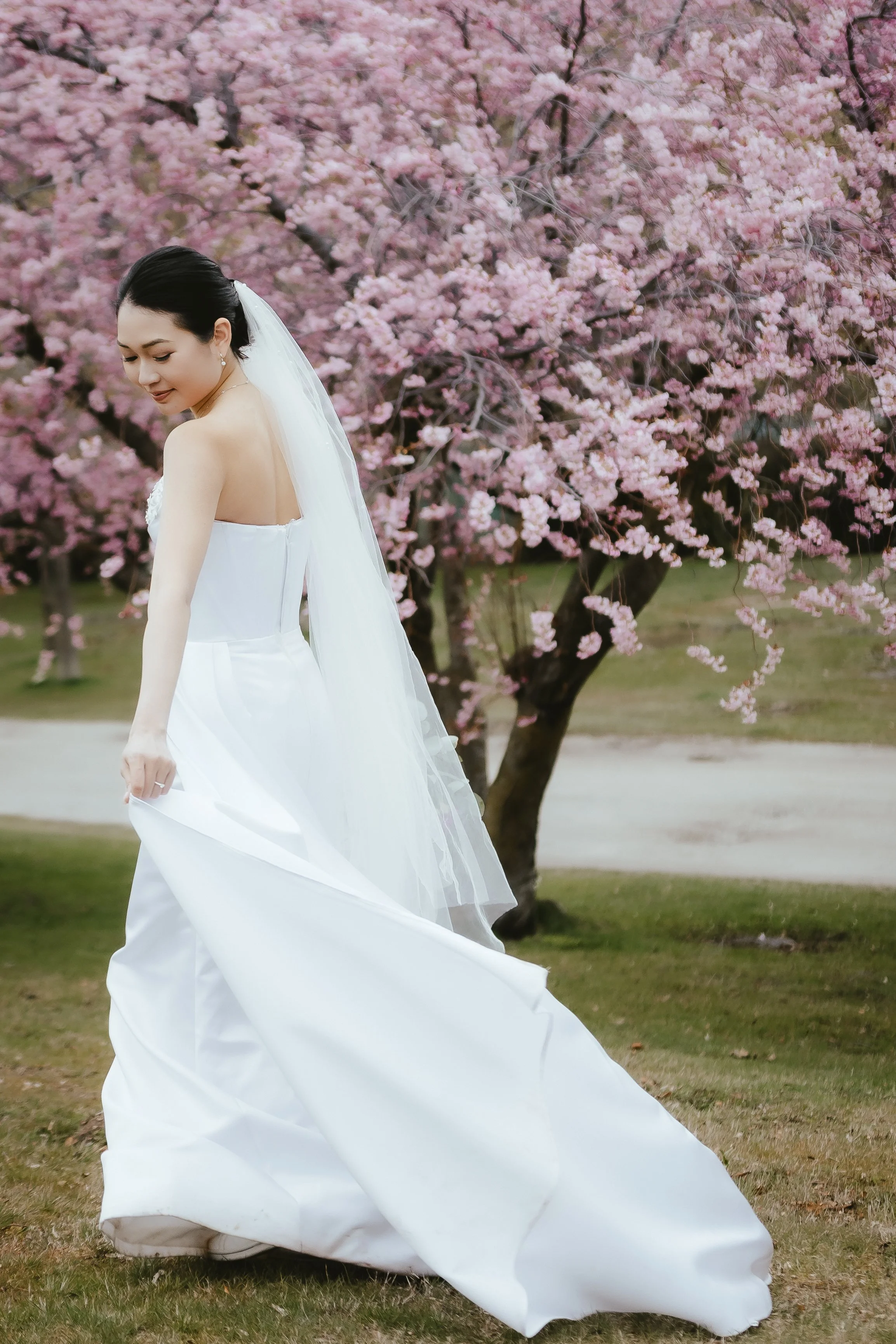 A woman in a white wedding dress and veil standing outdoors in front of pink cherry blossom trees.