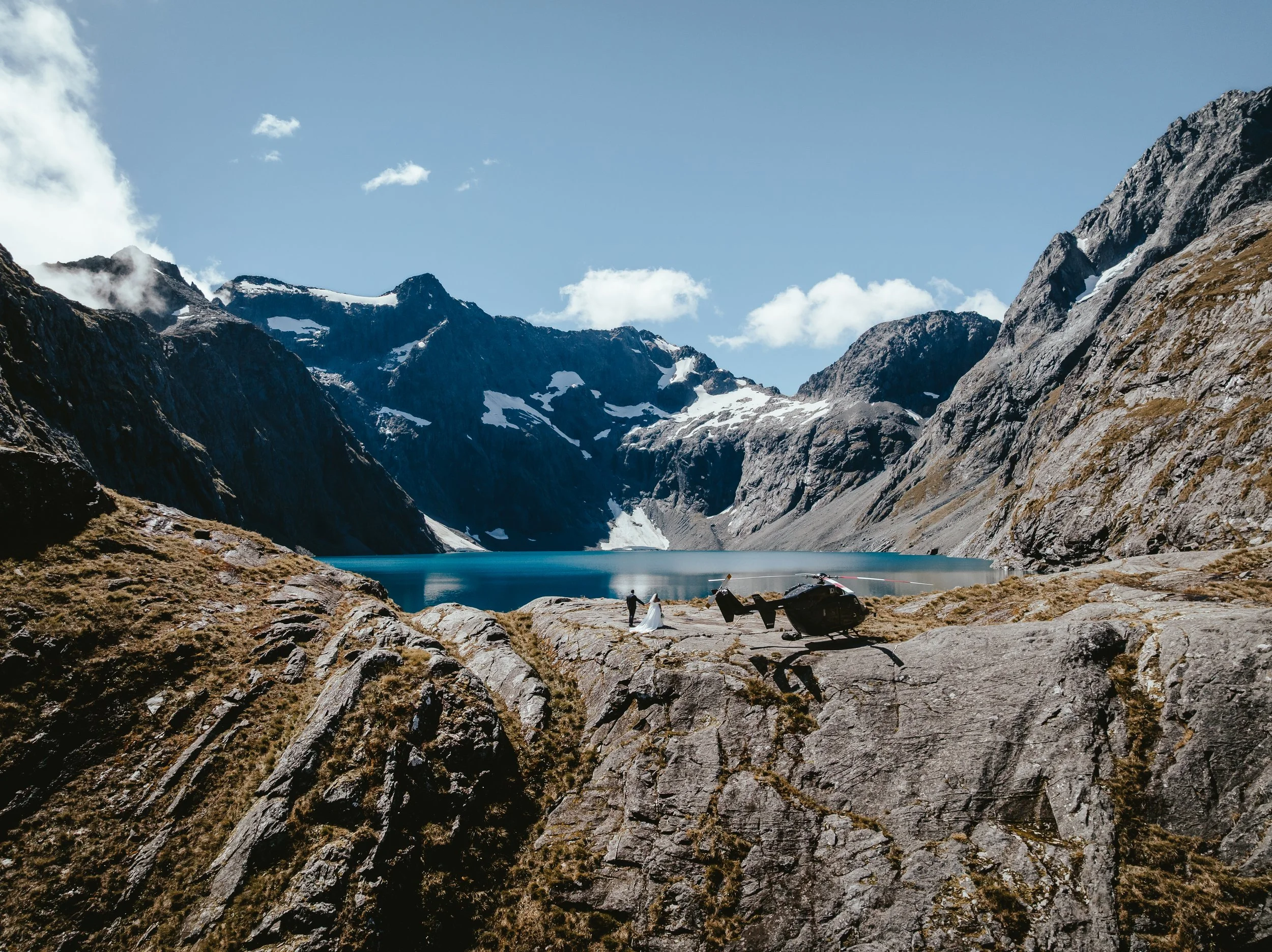 A scenic mountain landscape with snow-capped peaks, a mountain lake, and two people standing on rocky terrain near a helicopter.