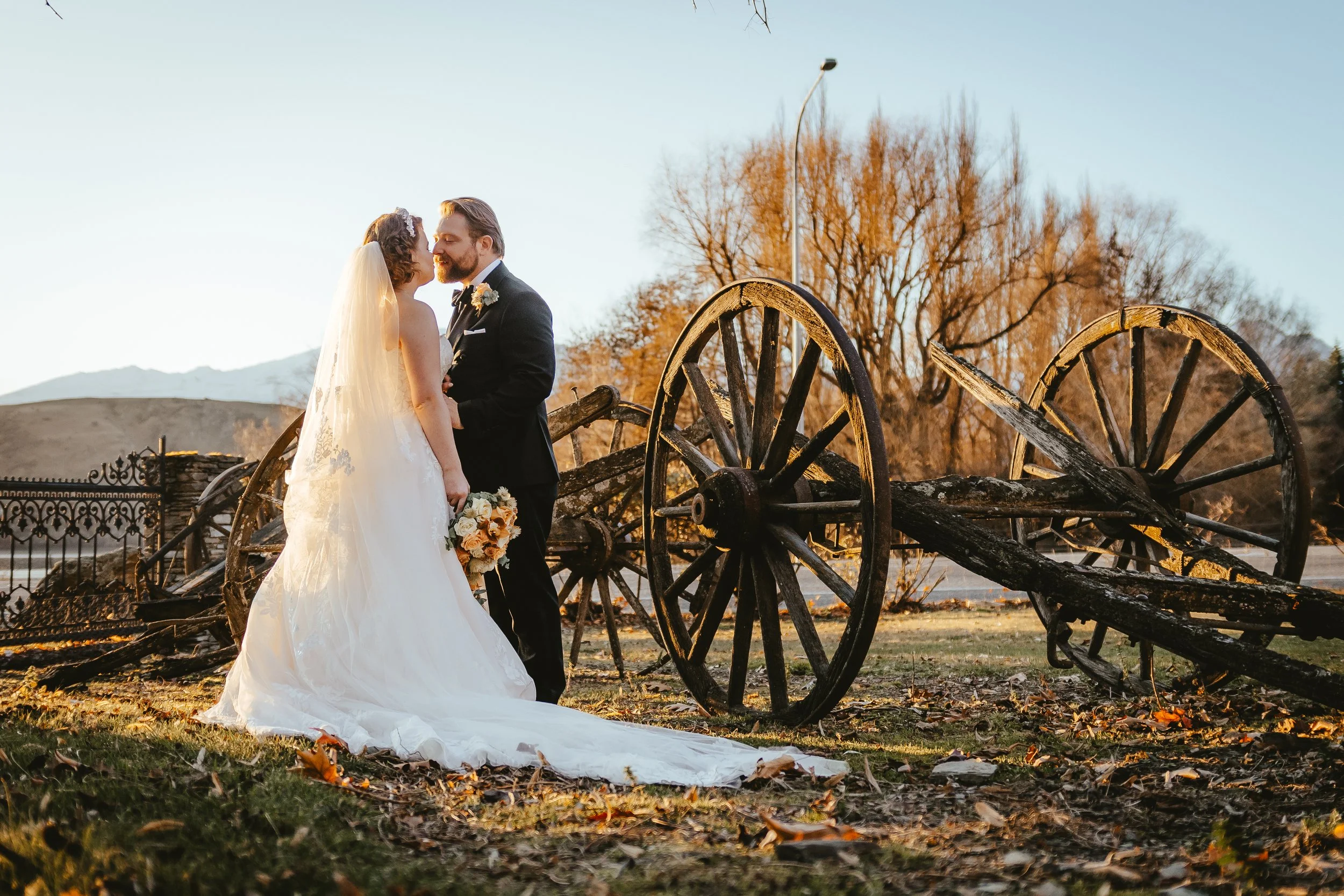 A bride and groom stand close together outdoors, sharing a moment on their wedding day near old wooden wagon wheels and in a rural setting during autumn.