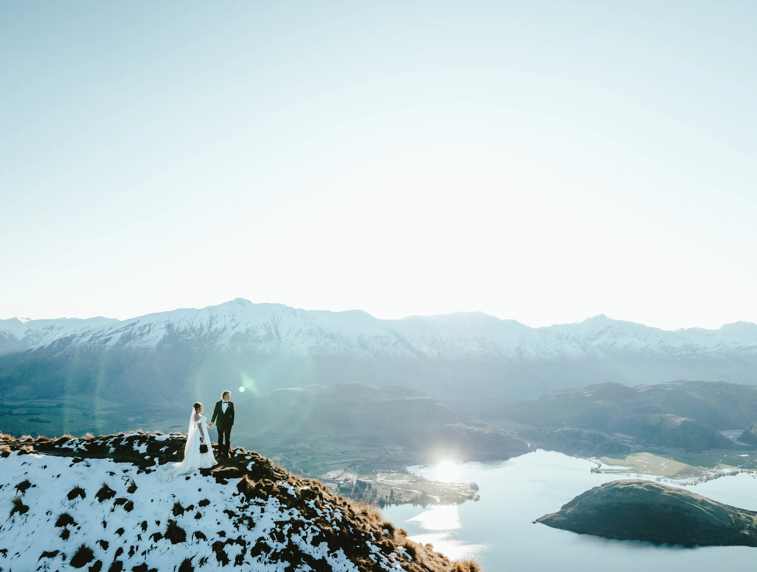 A bride and groom standing on snowy ground by a mountain lake with snow-capped mountains in the background, sunny sky, and reflection in the water.