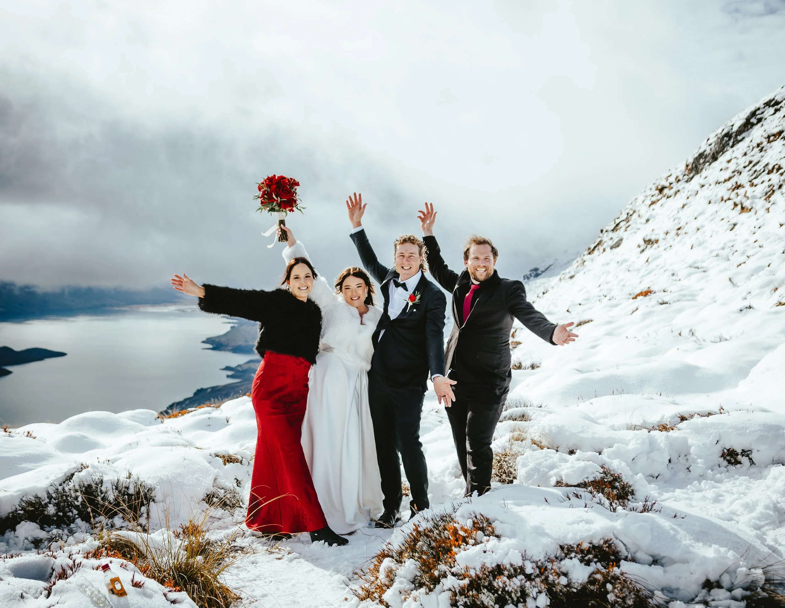 A group of seven people celebrating outdoors in a snowy mountainous area, with one person holding a bouquet of red roses. They are smiling and raising their arms in a joyful manner.