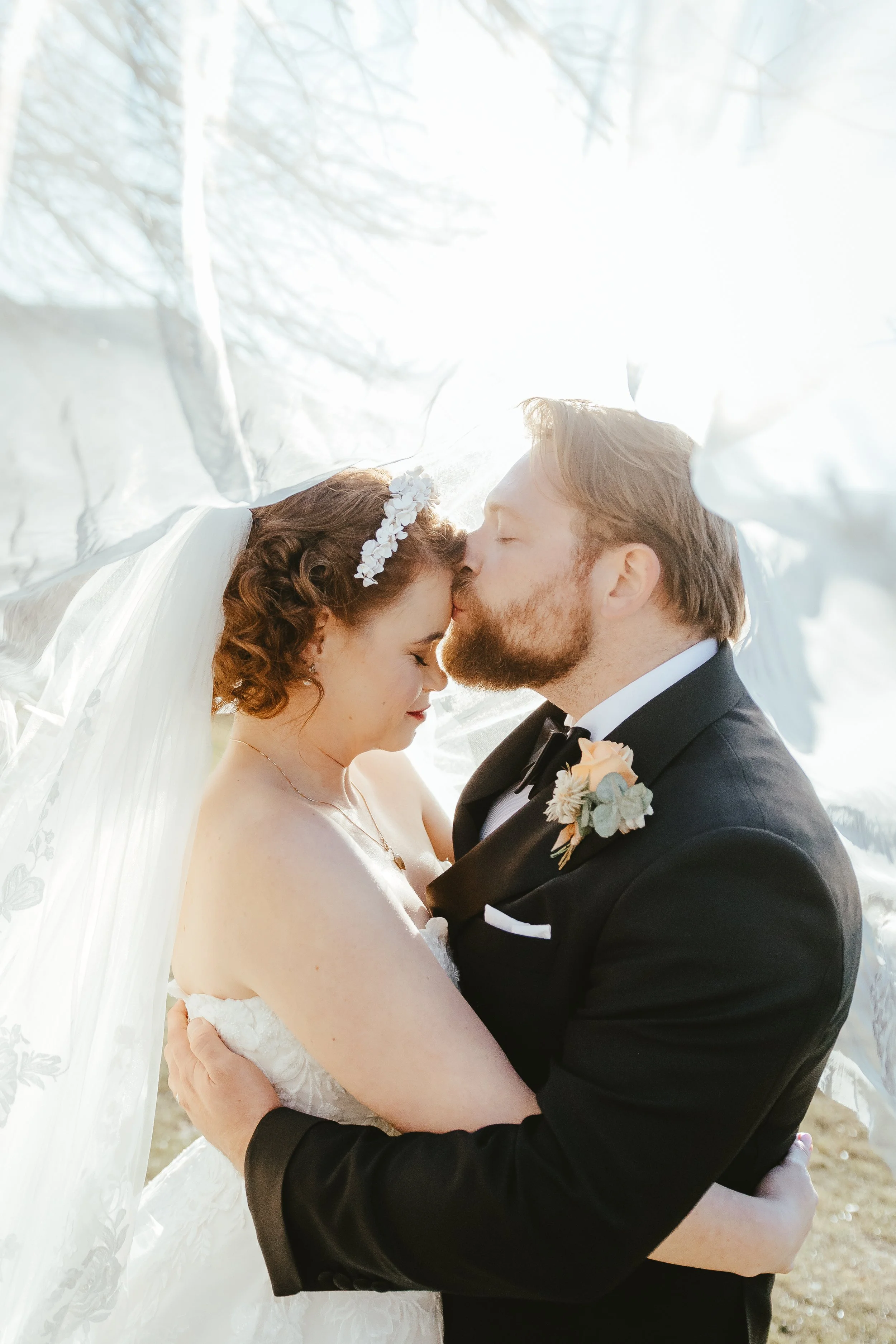 A bride and groom are sharing a tender moment, with their foreheads touching and eyes closed, under a white veil or fabric. The bride has curly hair adorned with a floral headpiece and is wearing a white wedding dress. The groom is in a black tuxedo 