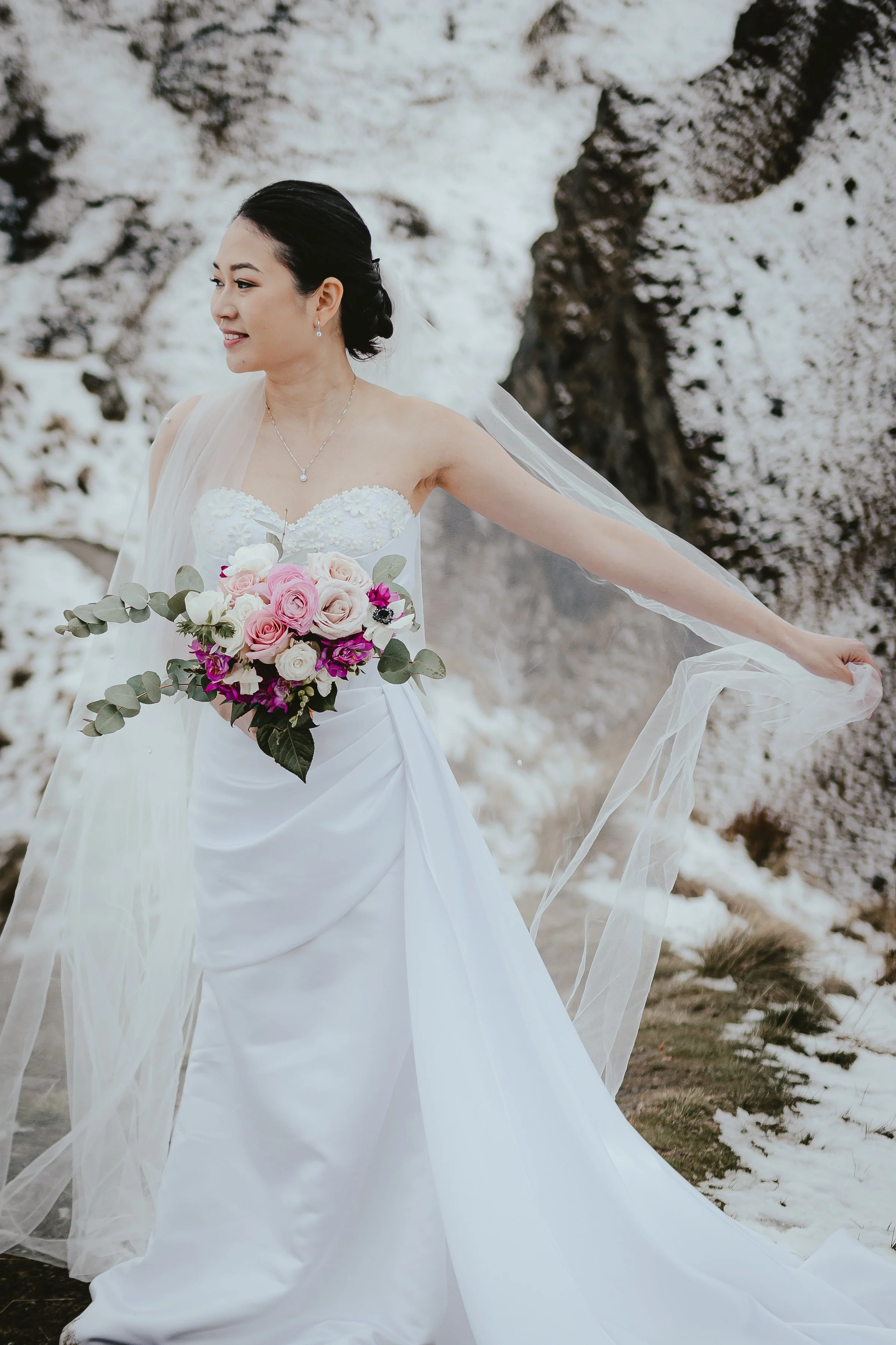 A bride in a white wedding gown holding a bouquet of pink, white, and purple flowers, standing outdoors in a snowy landscape.