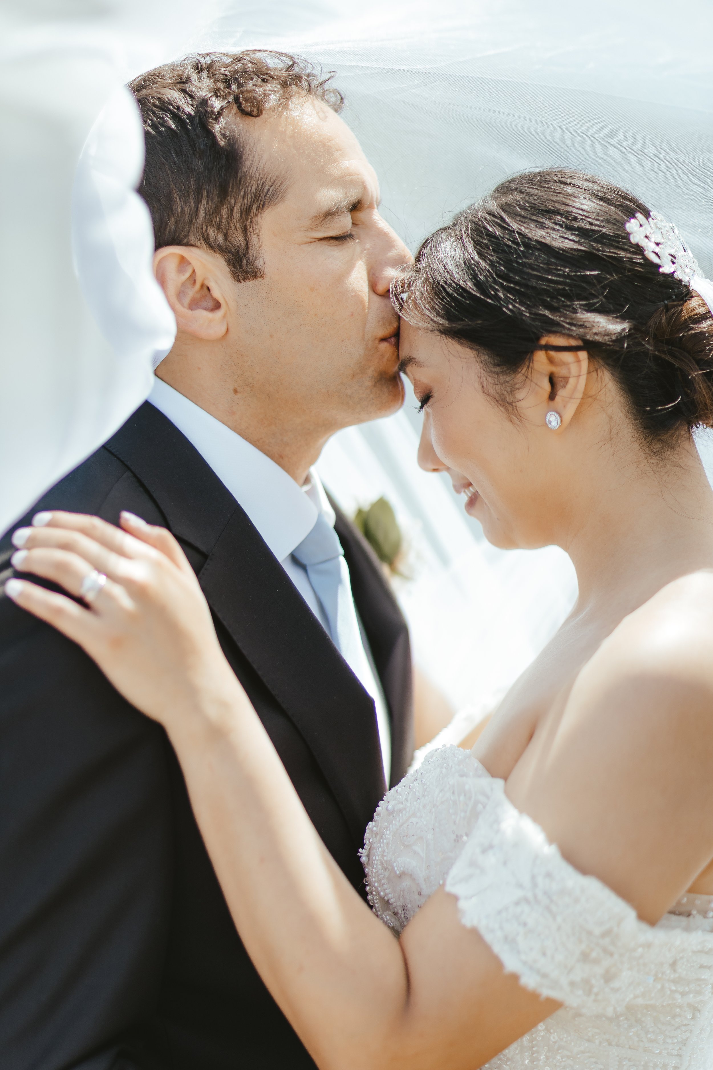 A bride and groom sharing a kiss during their wedding, with the groom in a black tuxedo and the bride in a white lace wedding dress, her hair adorned with a decorative hairpiece.