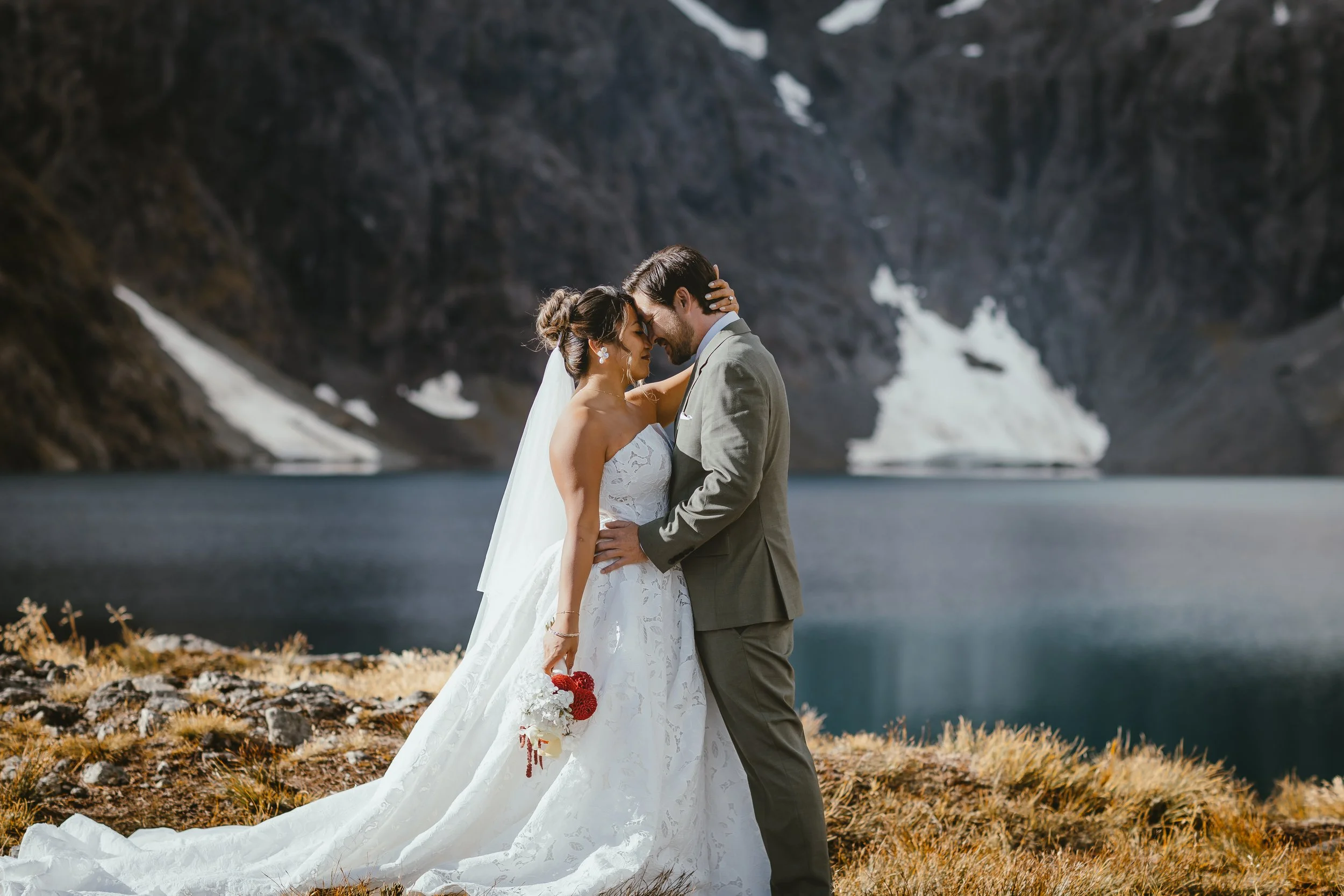 A bride and groom share a close, intimate moment outdoors near a lake with mountains and patches of snow in the background. The bride wears a white strapless wedding gown and the groom is in a gray suit. The bride holds a bouquet of red and white flo