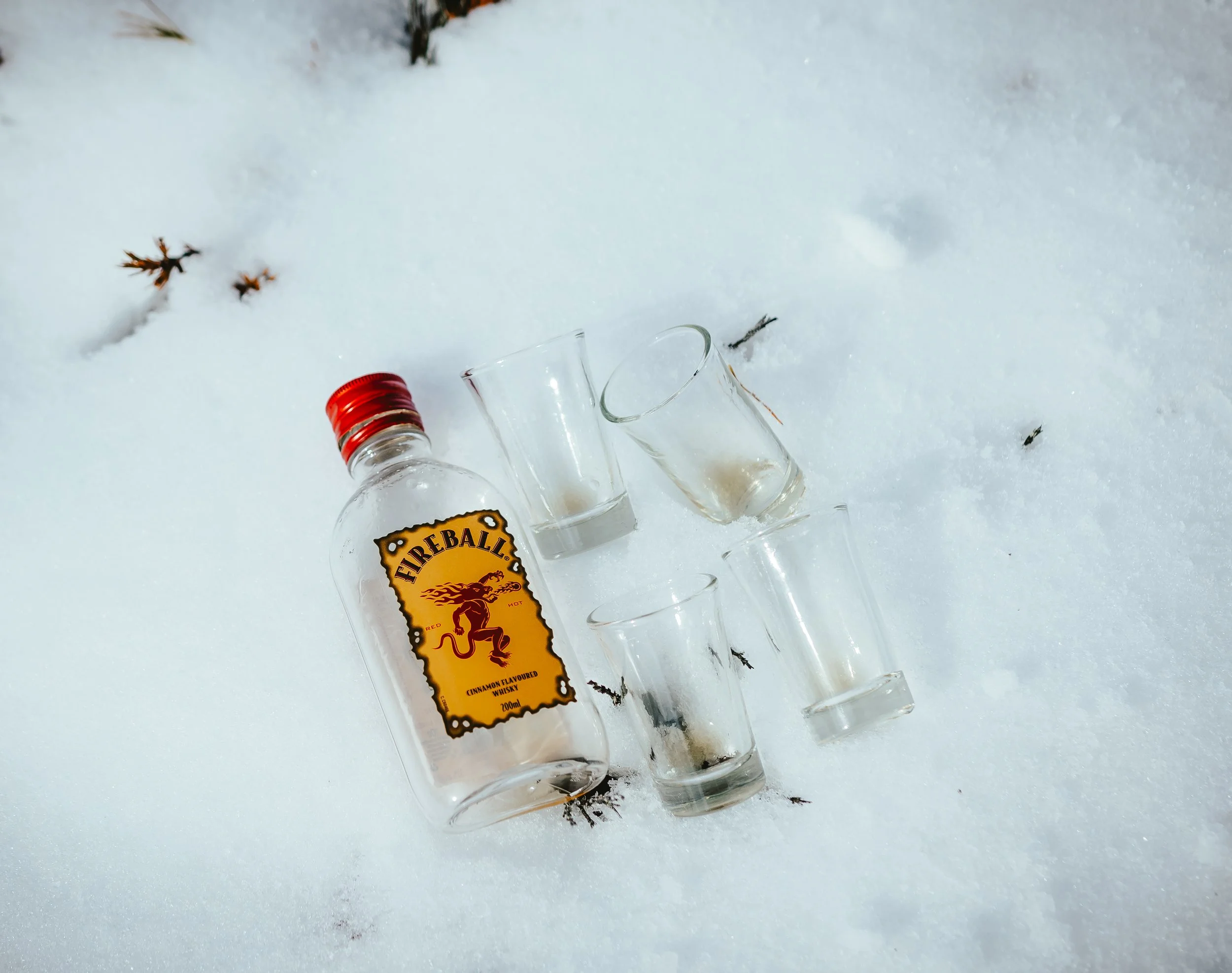 Several empty glass bottles and a glass on snow, with a visible bottle of cinnamon-flavored whiskey labeled 'Fireball' partially buried in snow.
