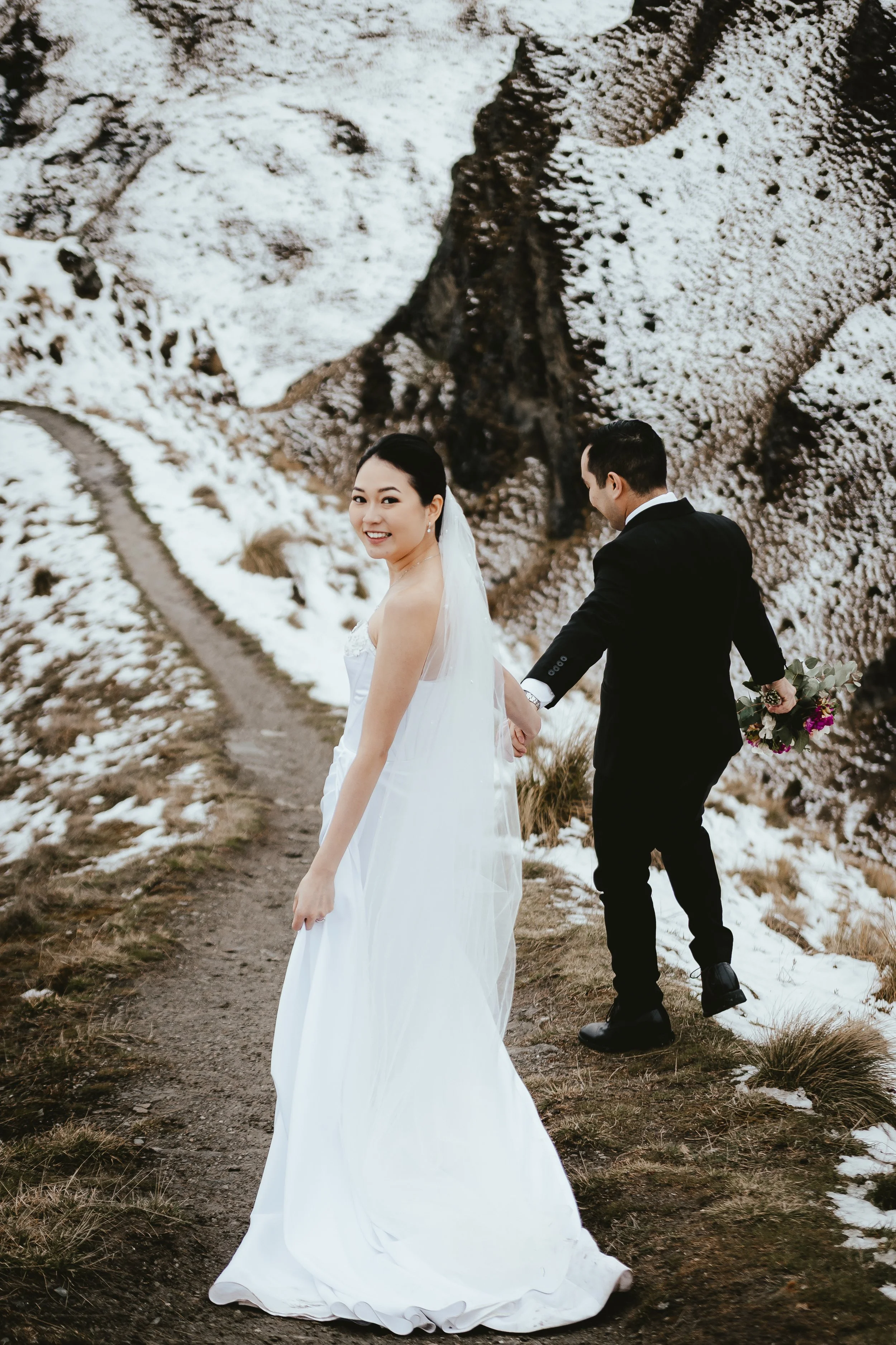A bride in a white wedding dress smiling at the camera and a groom in a black suit holding her hand while walking on a dirt path in a snowy outdoor setting.