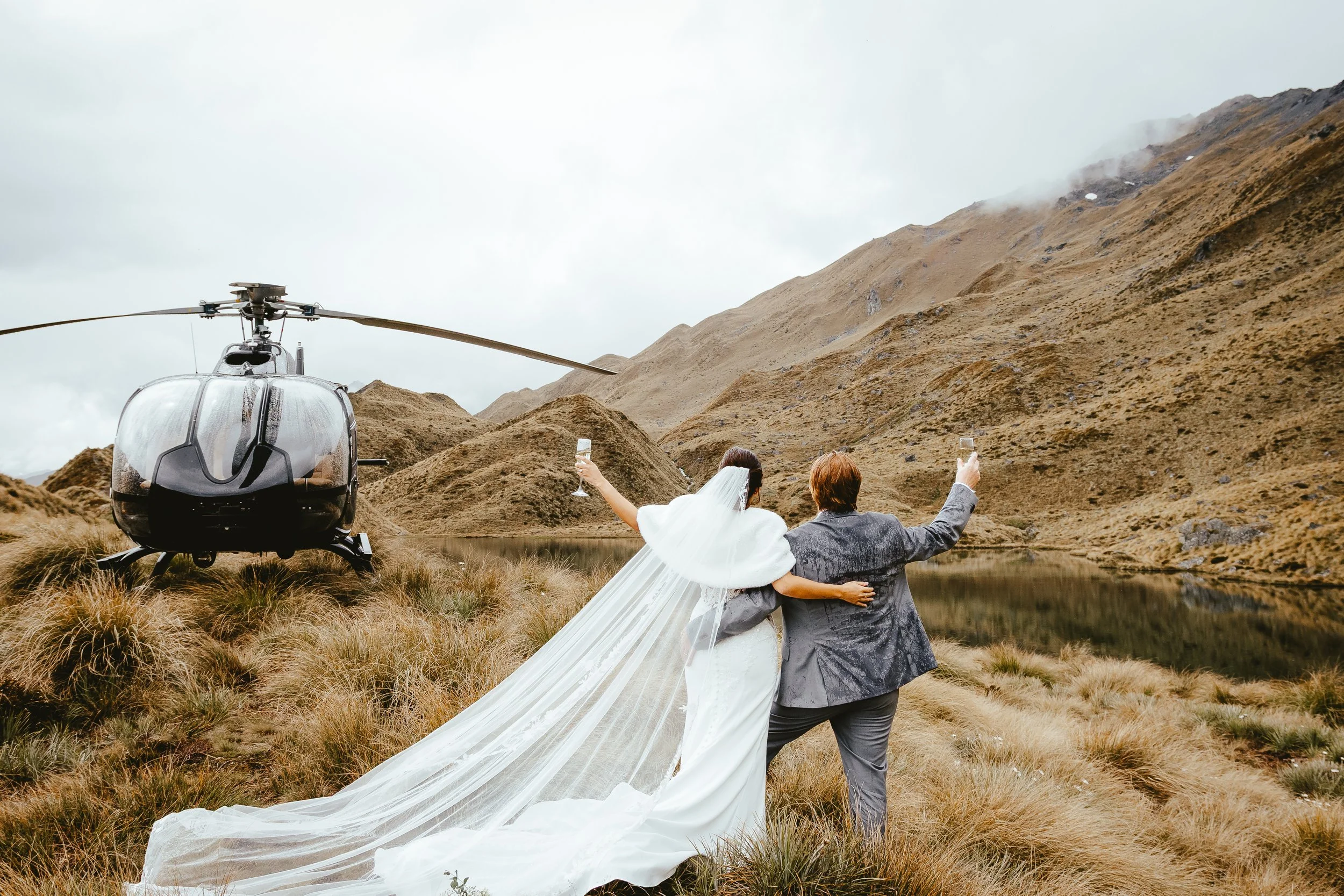 A bride and groom in wedding attire sitting on the grass beside a lake, holding glasses of champagne, with a helicopter parked nearby in a mountainous landscape.