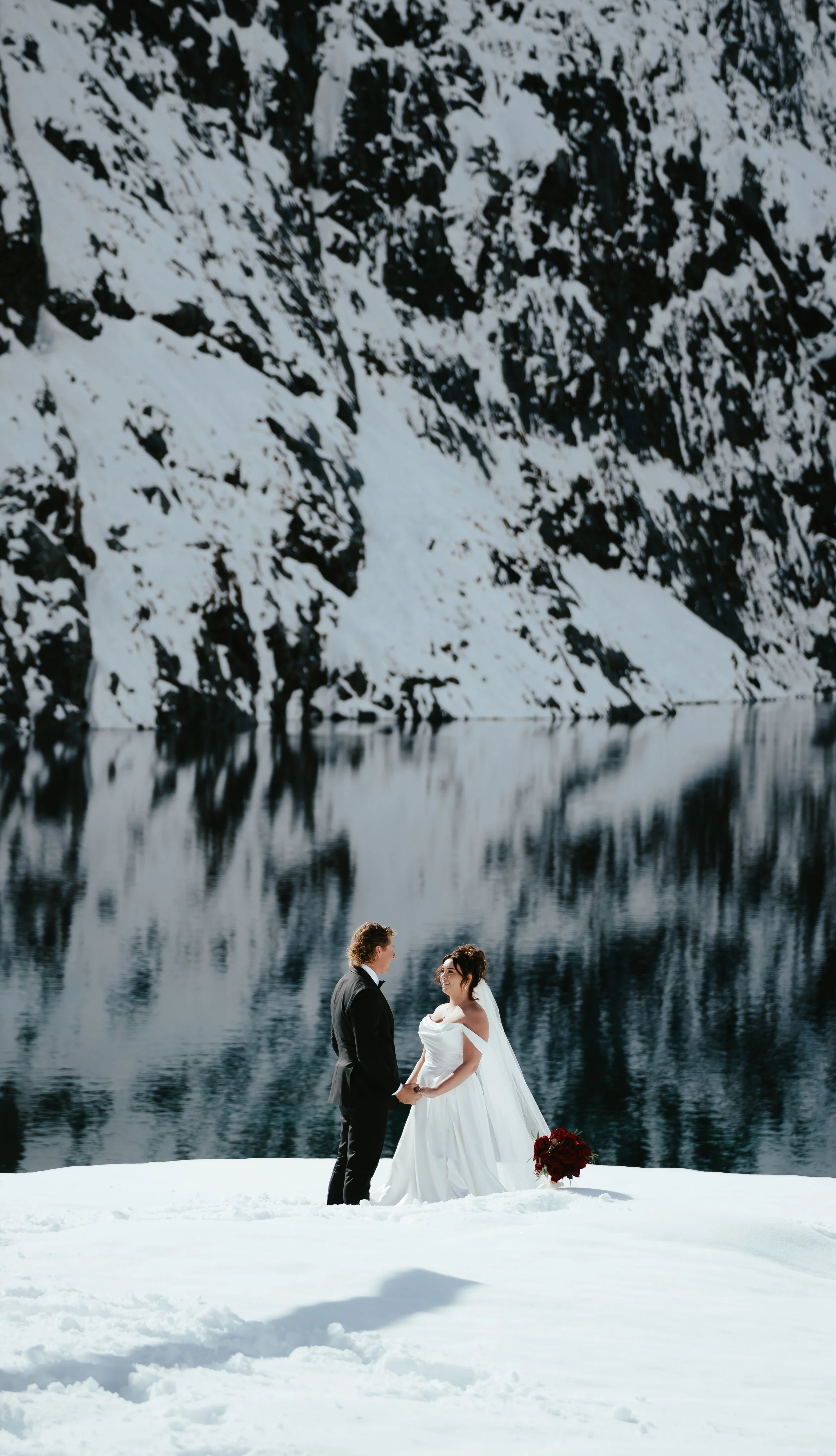 A bride and groom holding hands and facing each other on a snowy landscape beside a lake, with snow-covered mountains in the background.