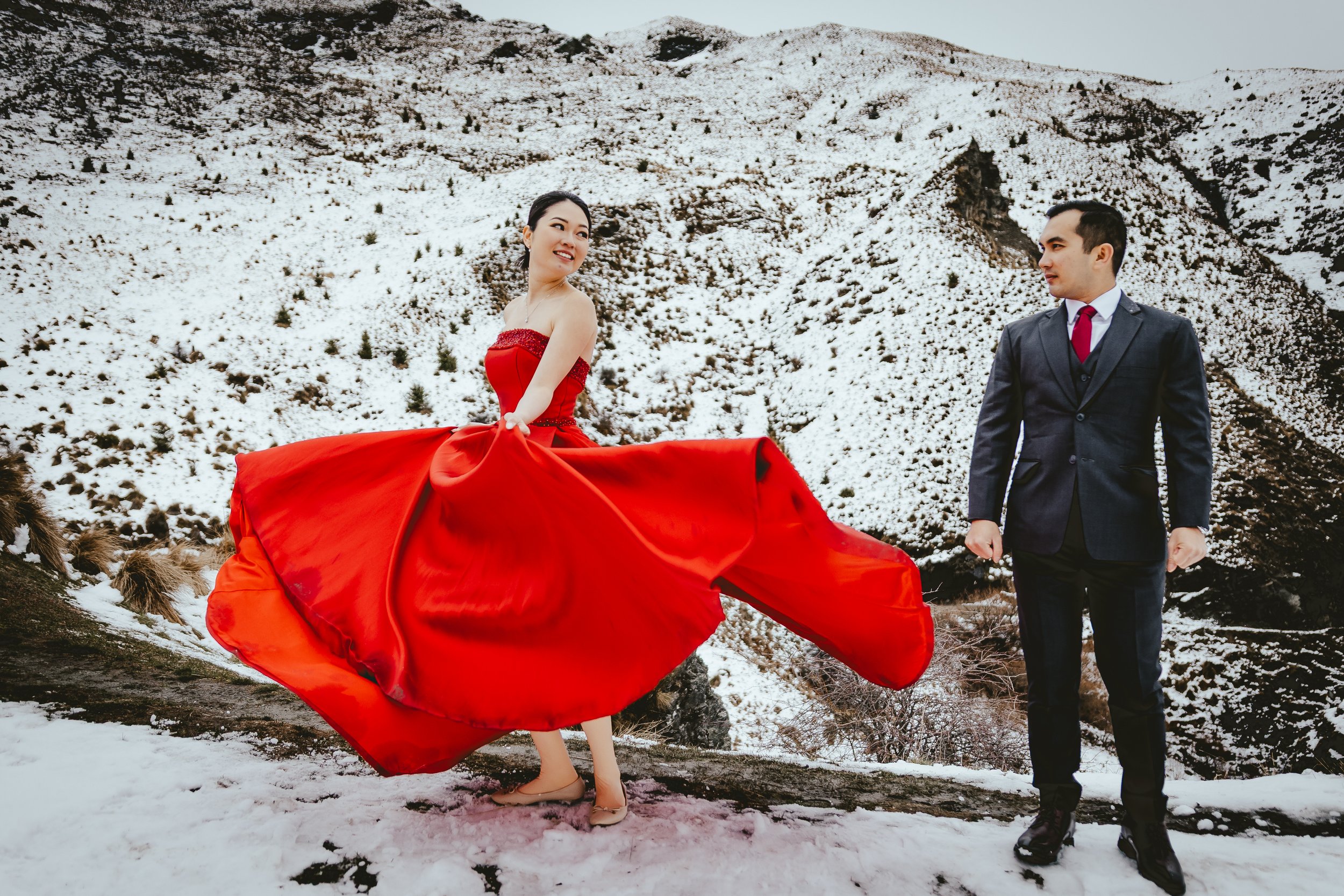 A woman in a long red dress and beige shoes is twirling in front of a snowy mountain landscape, while a man in a dark suit and red tie looks on.