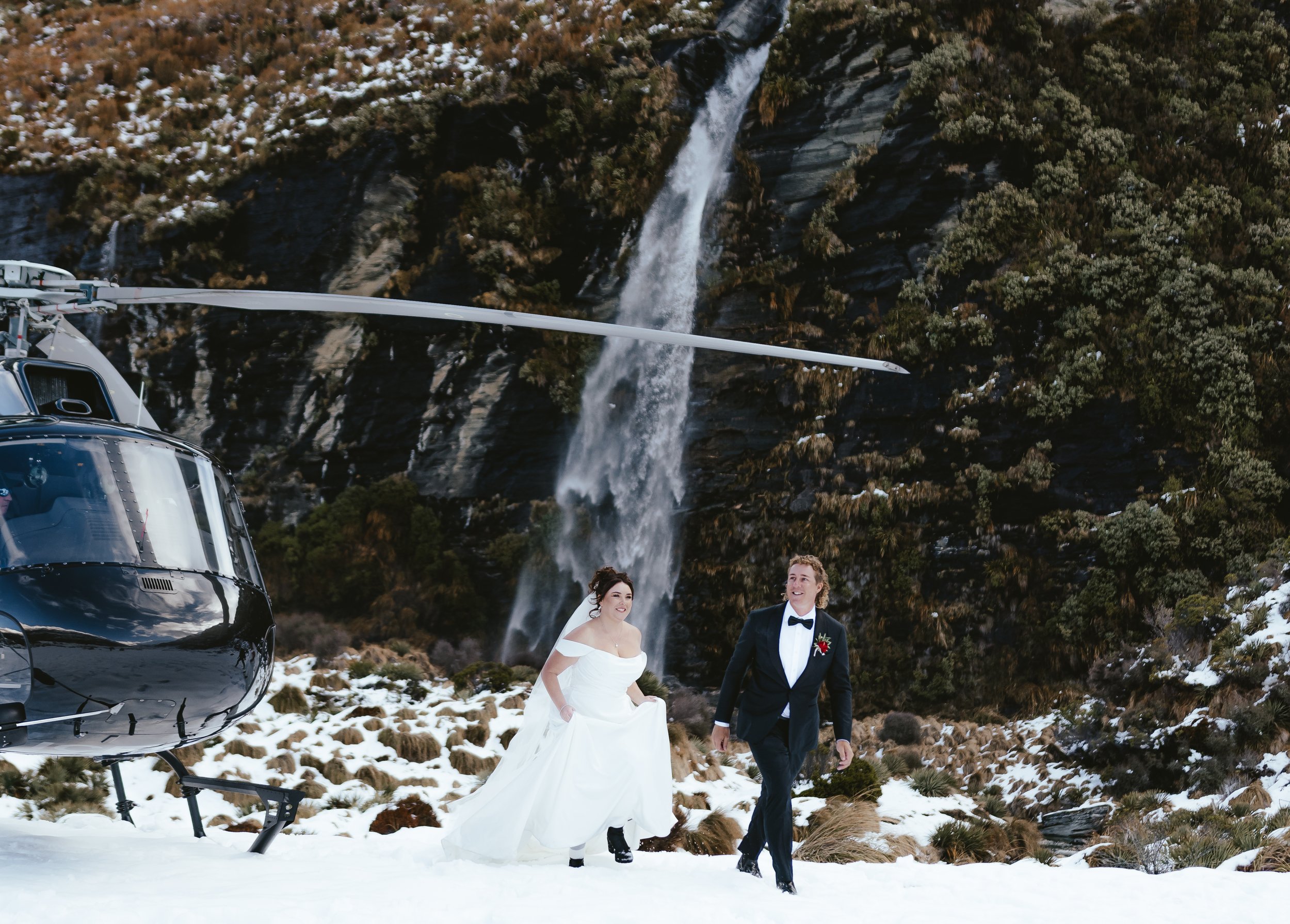 A newlywed couple in wedding attire walking in a snowy, mountainous landscape with a waterfall in the background. A helicopter is partially visible on the left side of the image.