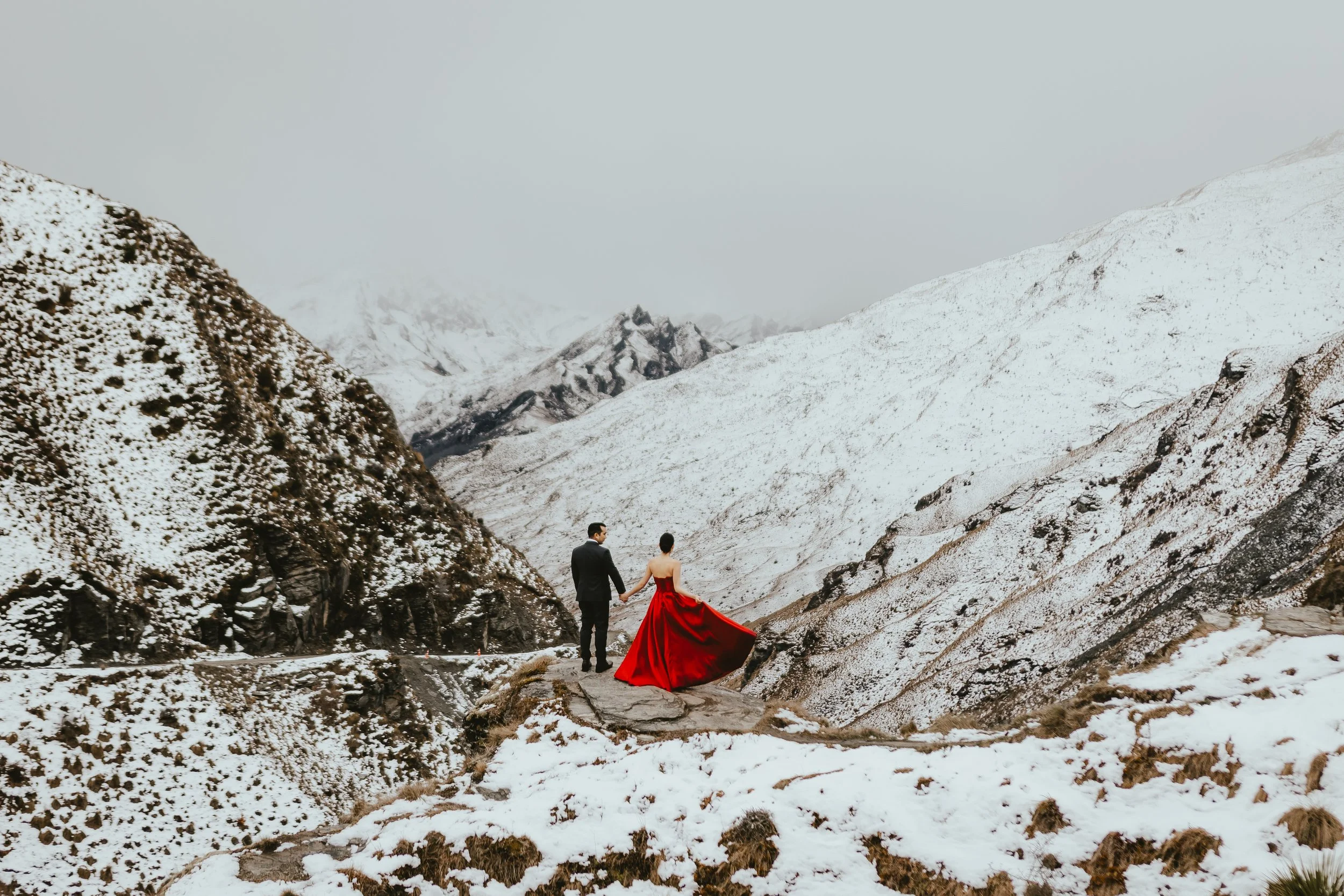 A couple holding hands in a snowy mountain landscape, with one person wearing a red dress and the other in formal attire.