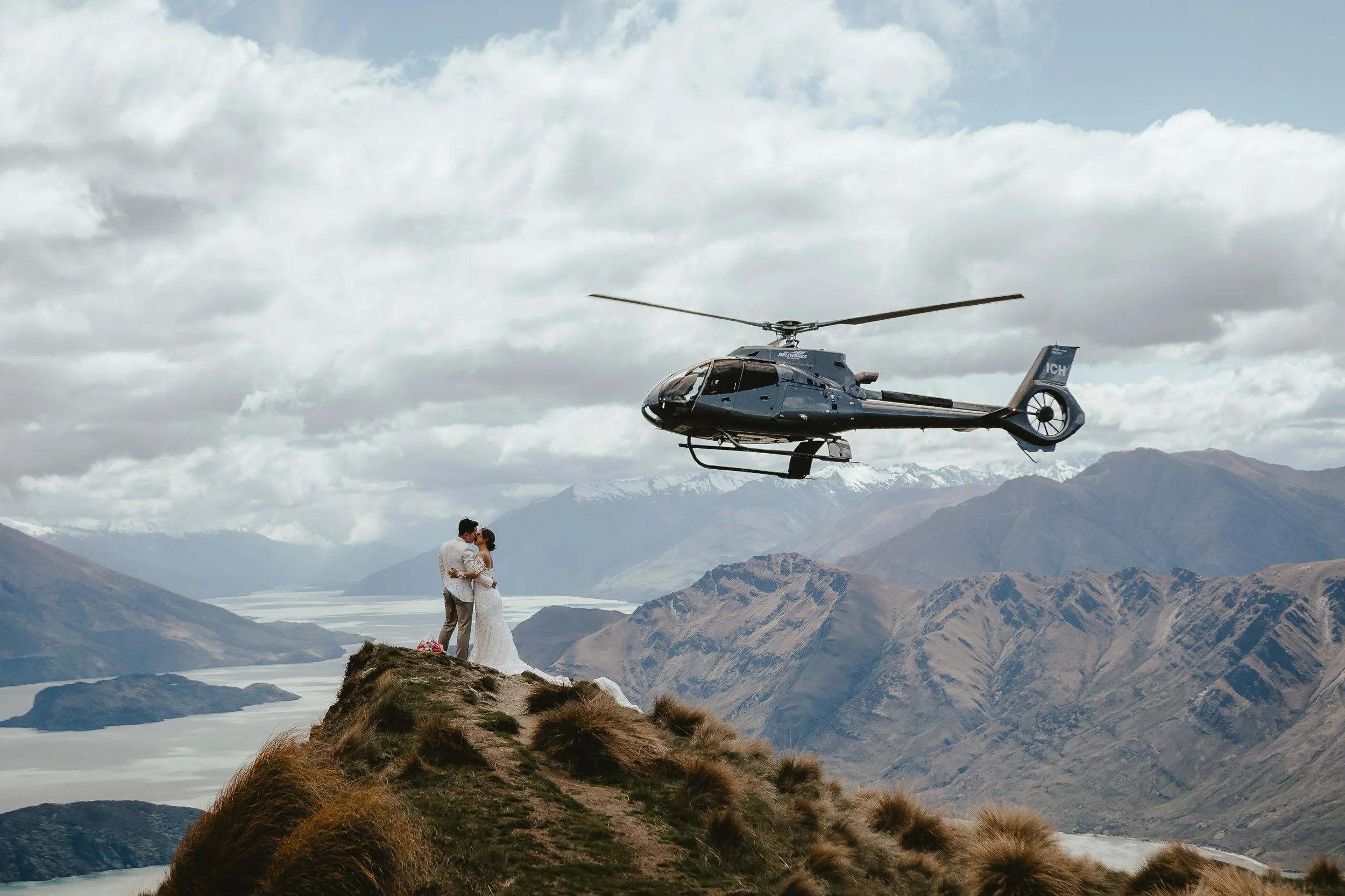 A couple dressed in wedding attire standing on a mountain ridge, with a helicopter flying overhead and a scenic landscape of mountains and water in the background.