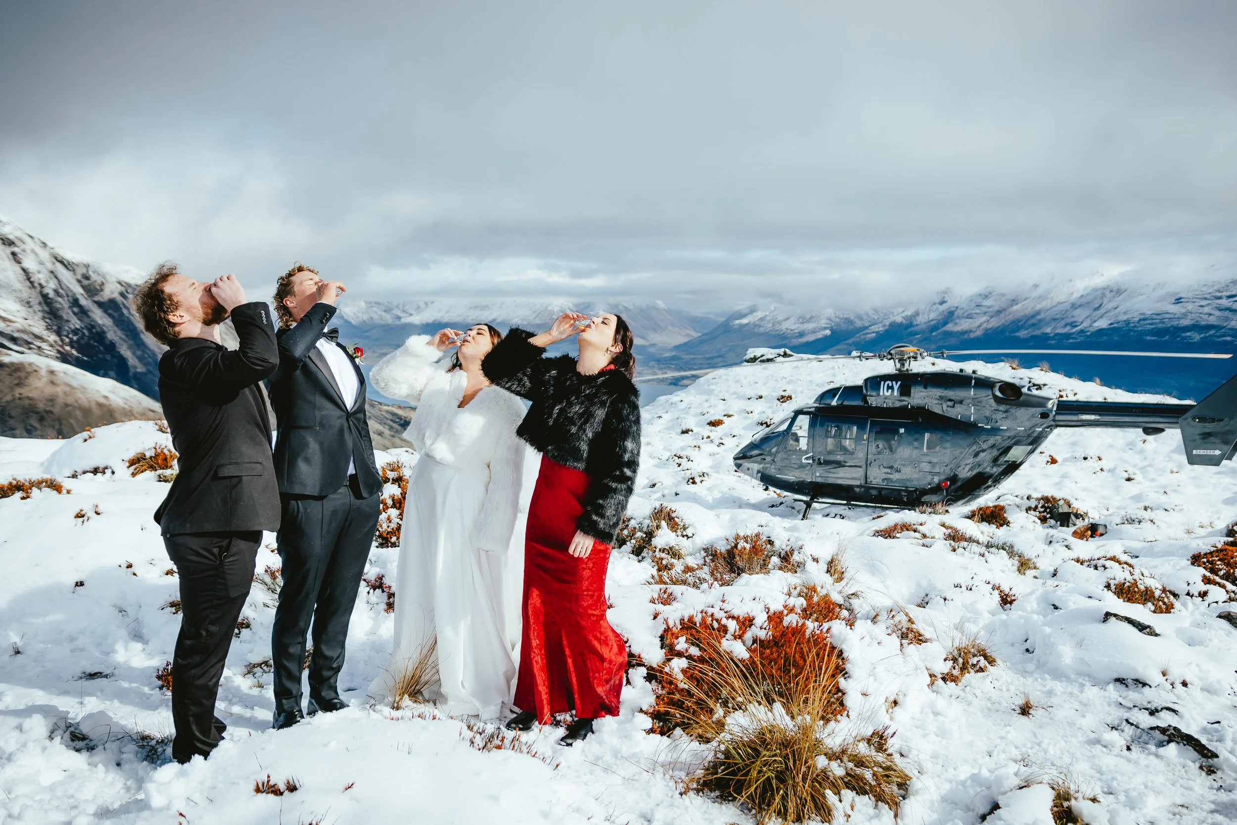 Four people in formal wedding attire standing on snow-covered ground in a mountainous outdoor setting, drinking from glasses, with a helicopter in the background.