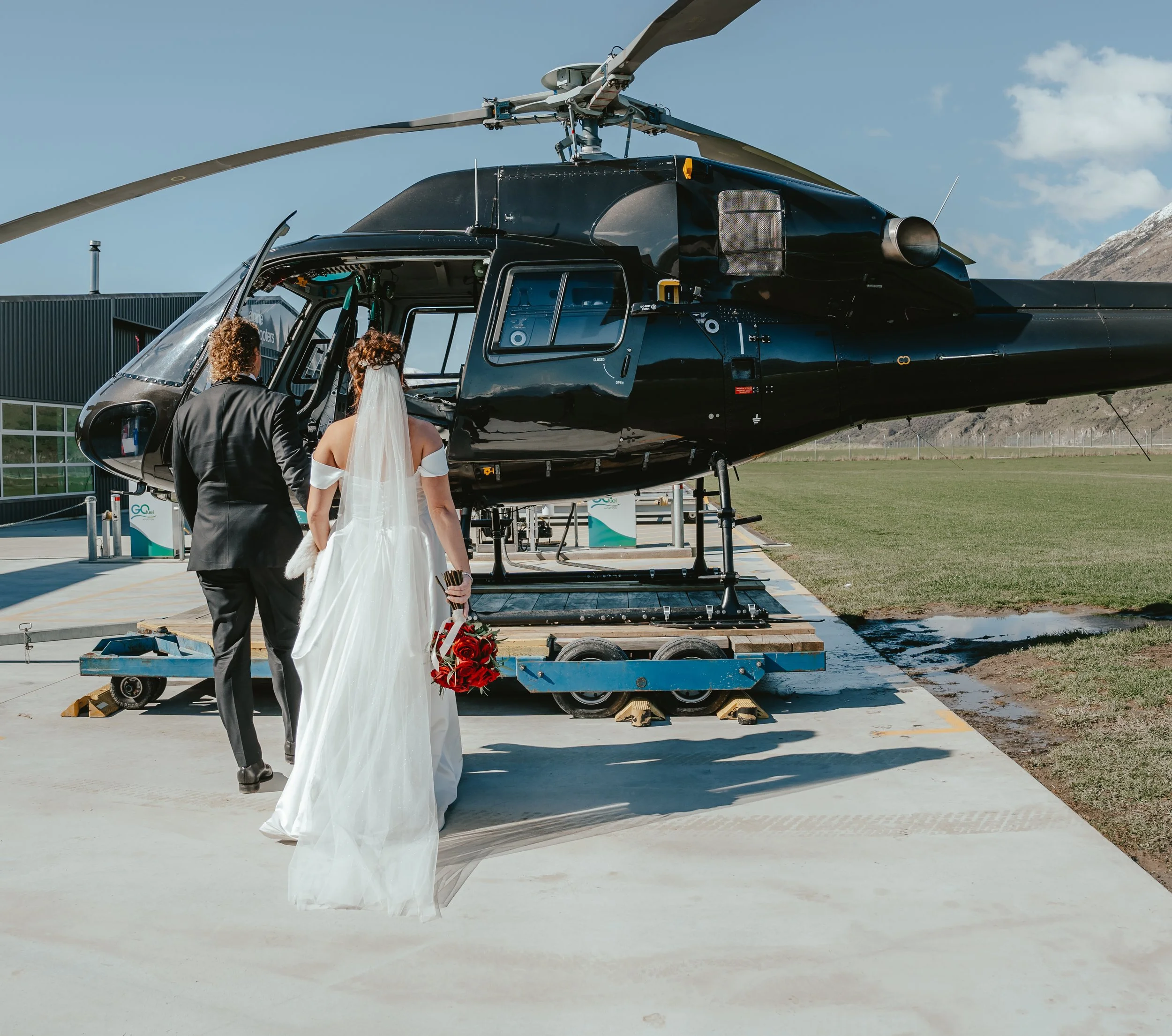 A bride and groom stand next to a black helicopter on a tarmac, with a bouquet of red roses on the ground nearby.