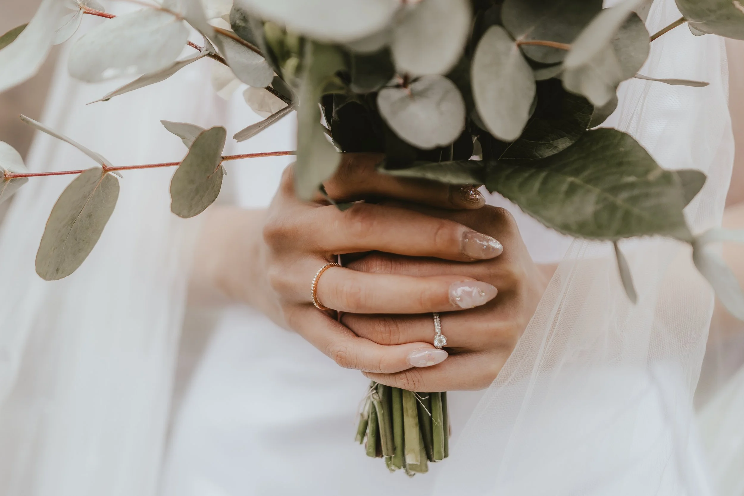 Person holding a bouquet of green and white eucalyptus leaves and stems, wearing rings and a white outfit.