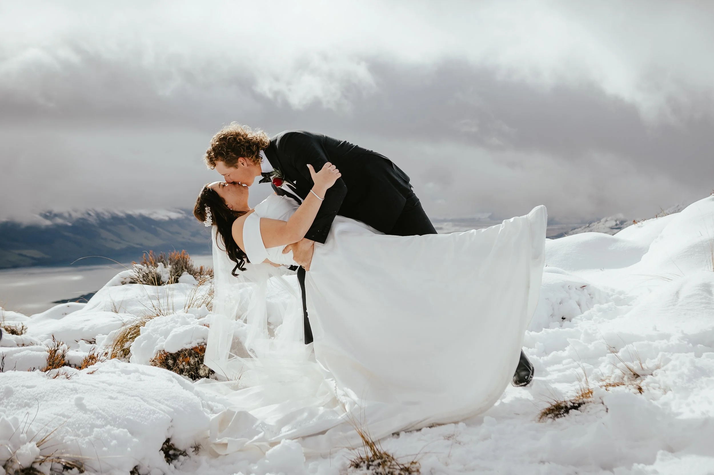 A couple in wedding attire kissing in a snowy outdoor landscape, with overcast skies and distant mountains.