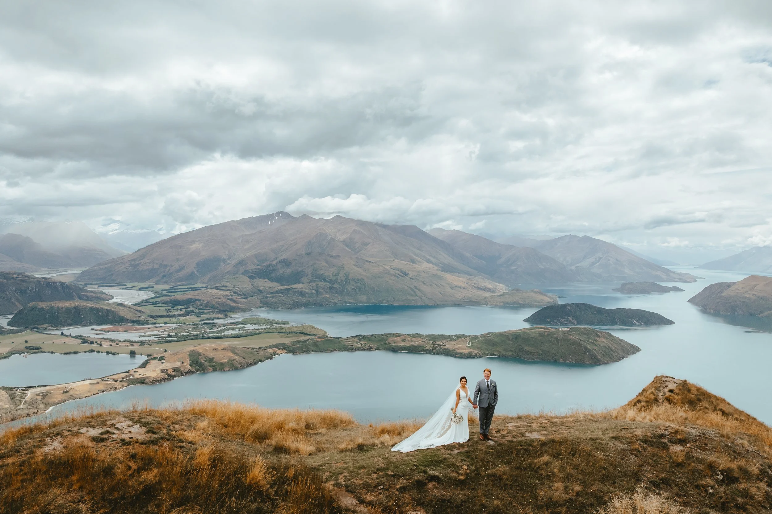 A newlywed couple standing on a hill with a scenic lake and mountain landscape in the background, under a cloudy sky.