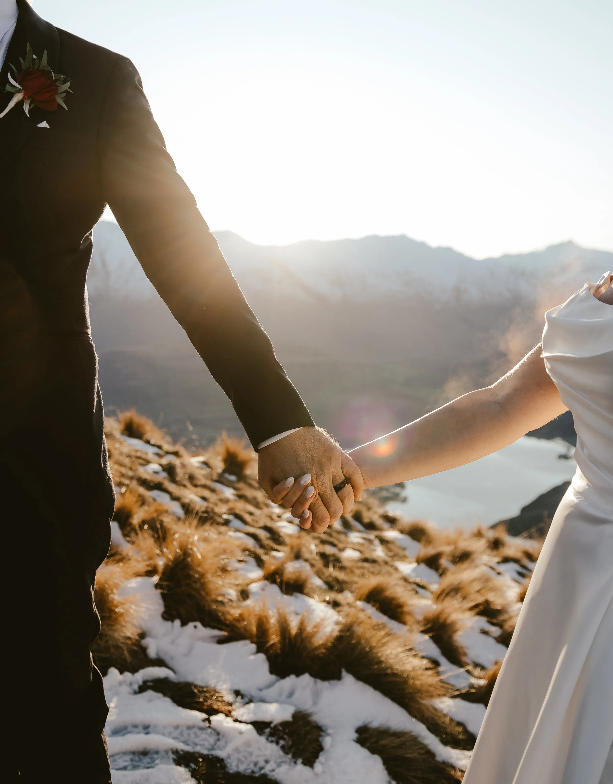Close-up of two people holding hands outdoors in a mountainous area with snow and dry grass, with bright sunlight and a clear sky.