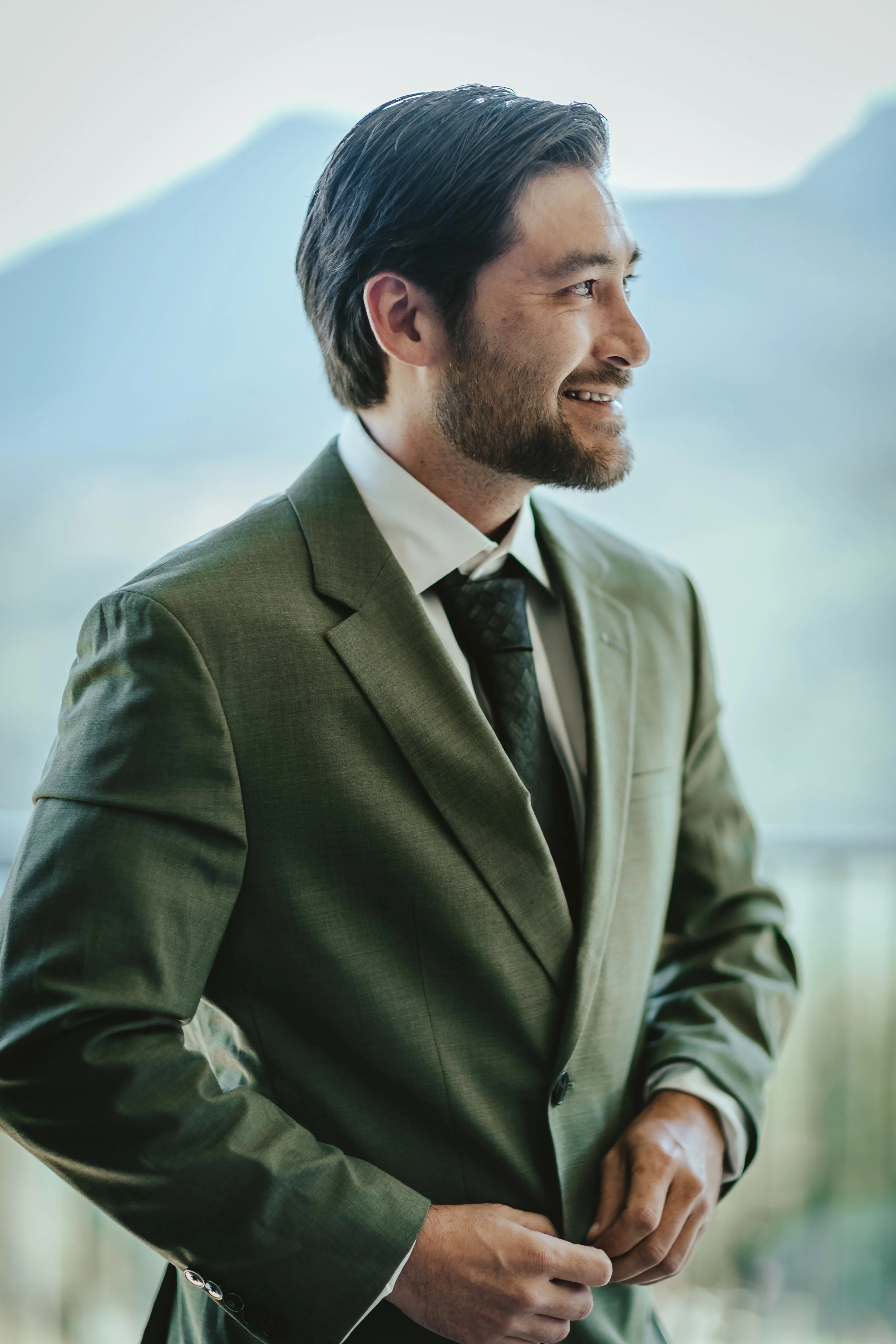 A man in a suit smiling and looking to the right, with mountains in the background.
