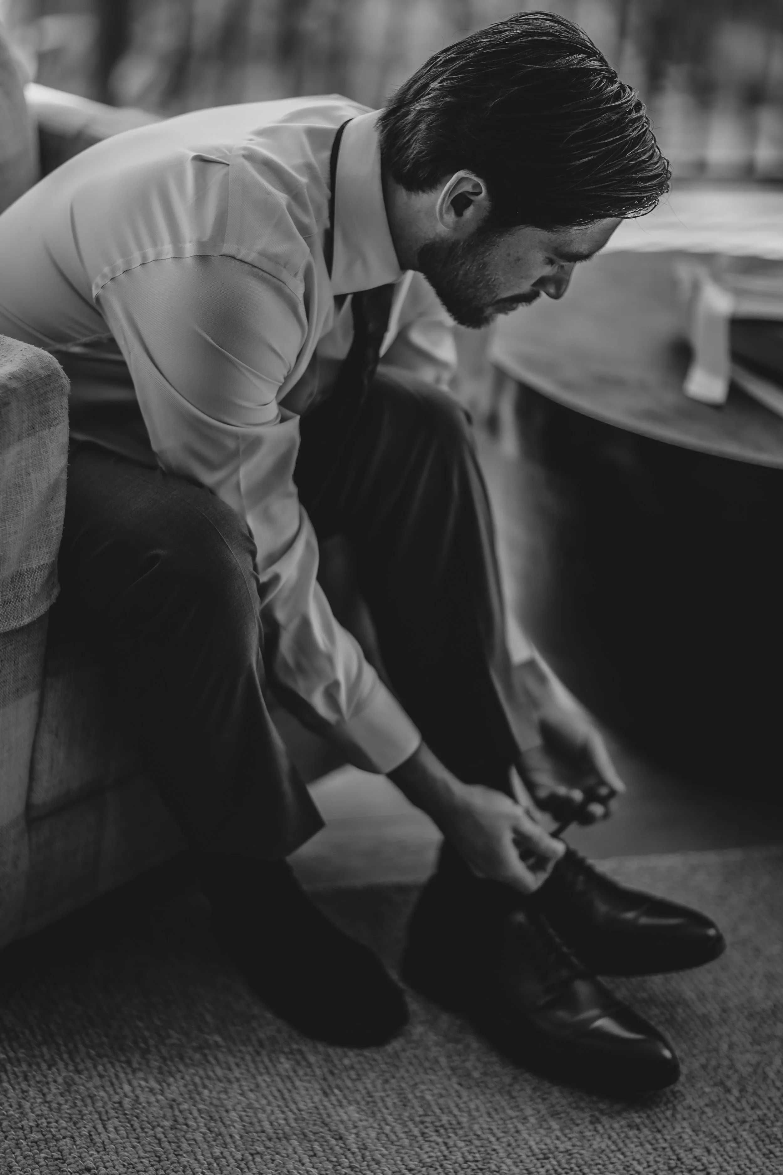 A man in formal attire is sitting on a sofa, leaning forward to put on dress shoes.