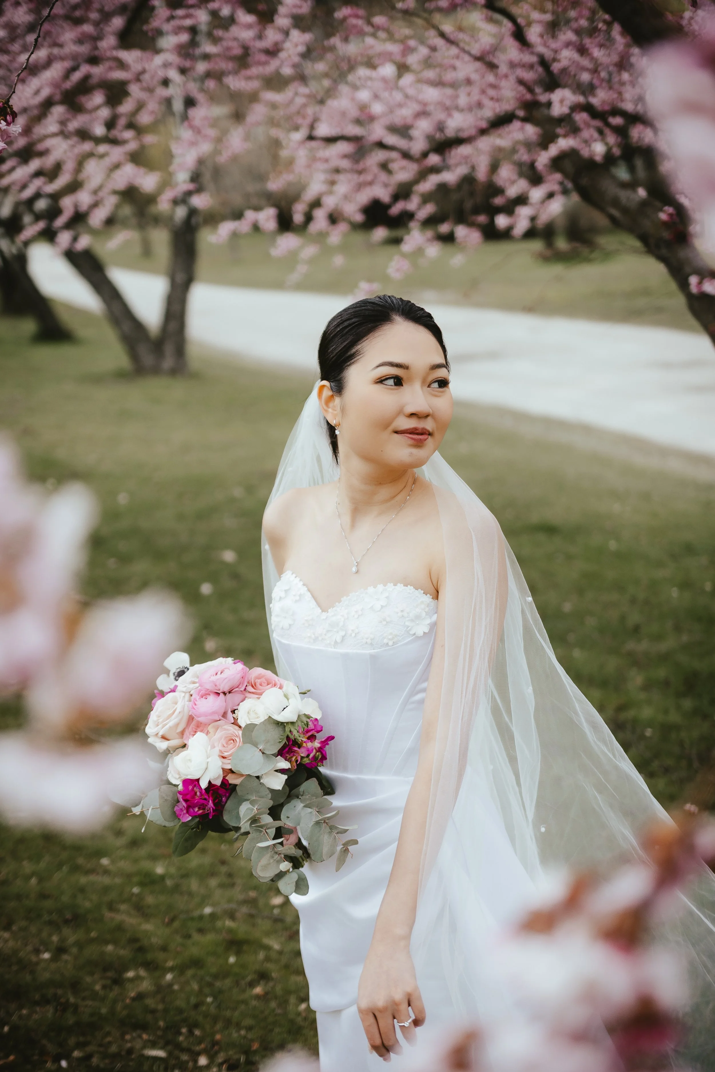 A bride in a white wedding dress holding a bouquet of pink, white, and purple flowers, standing outdoors beneath cherry blossom trees with pink flowers.
