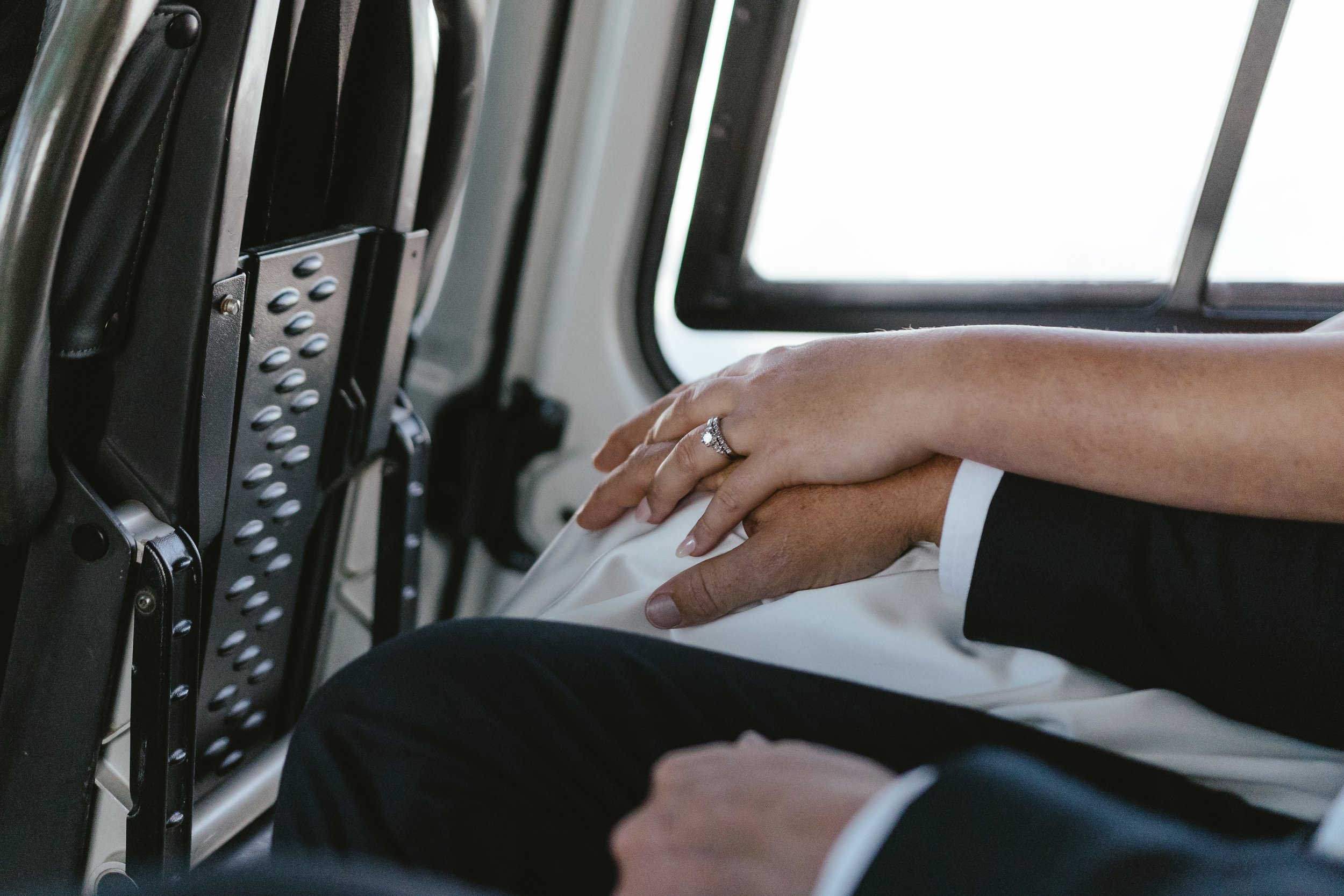 A close-up view of two people holding hands inside an airplane, with one person wearing a wedding ring. The background shows airplane window and seat controls.