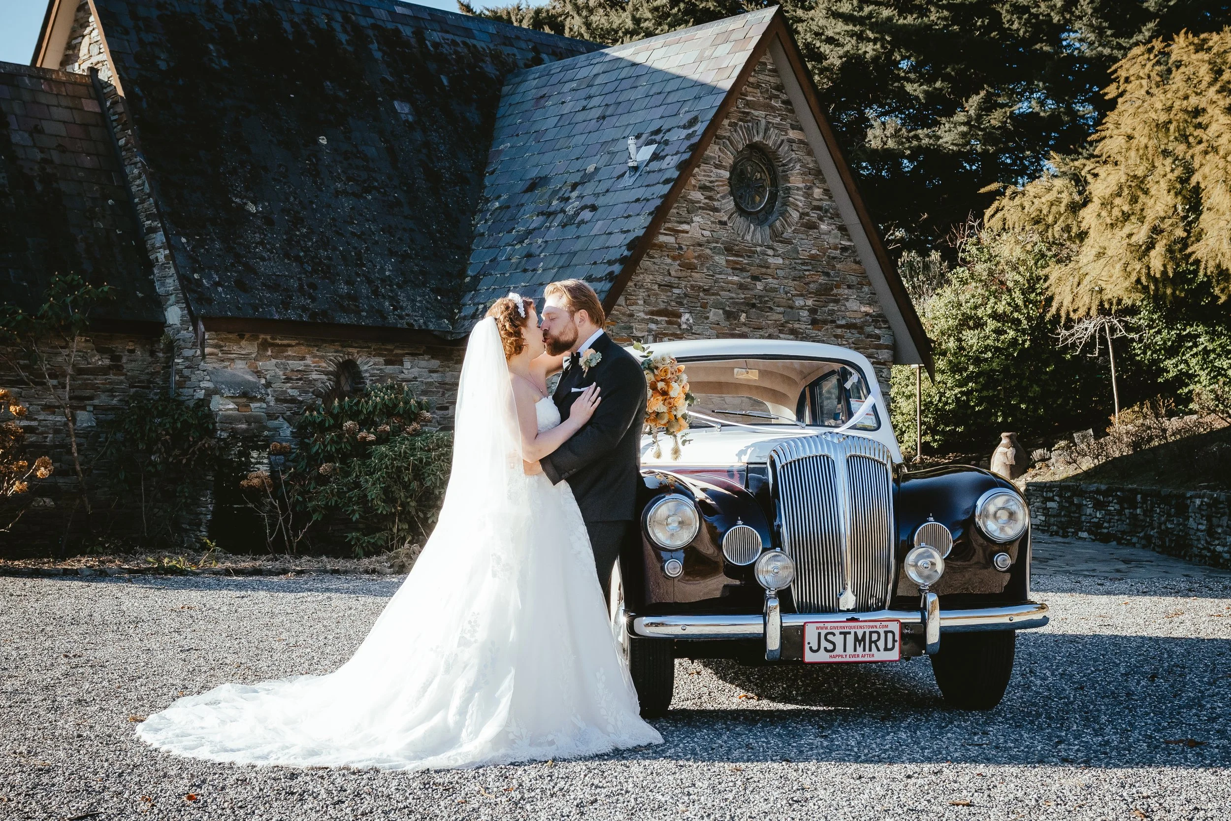 A newlywed couple in wedding attire kissing in front of a vintage black car with a floral bouquet on the side, parked in front of a stone church with a steeple, under a clear sky.
