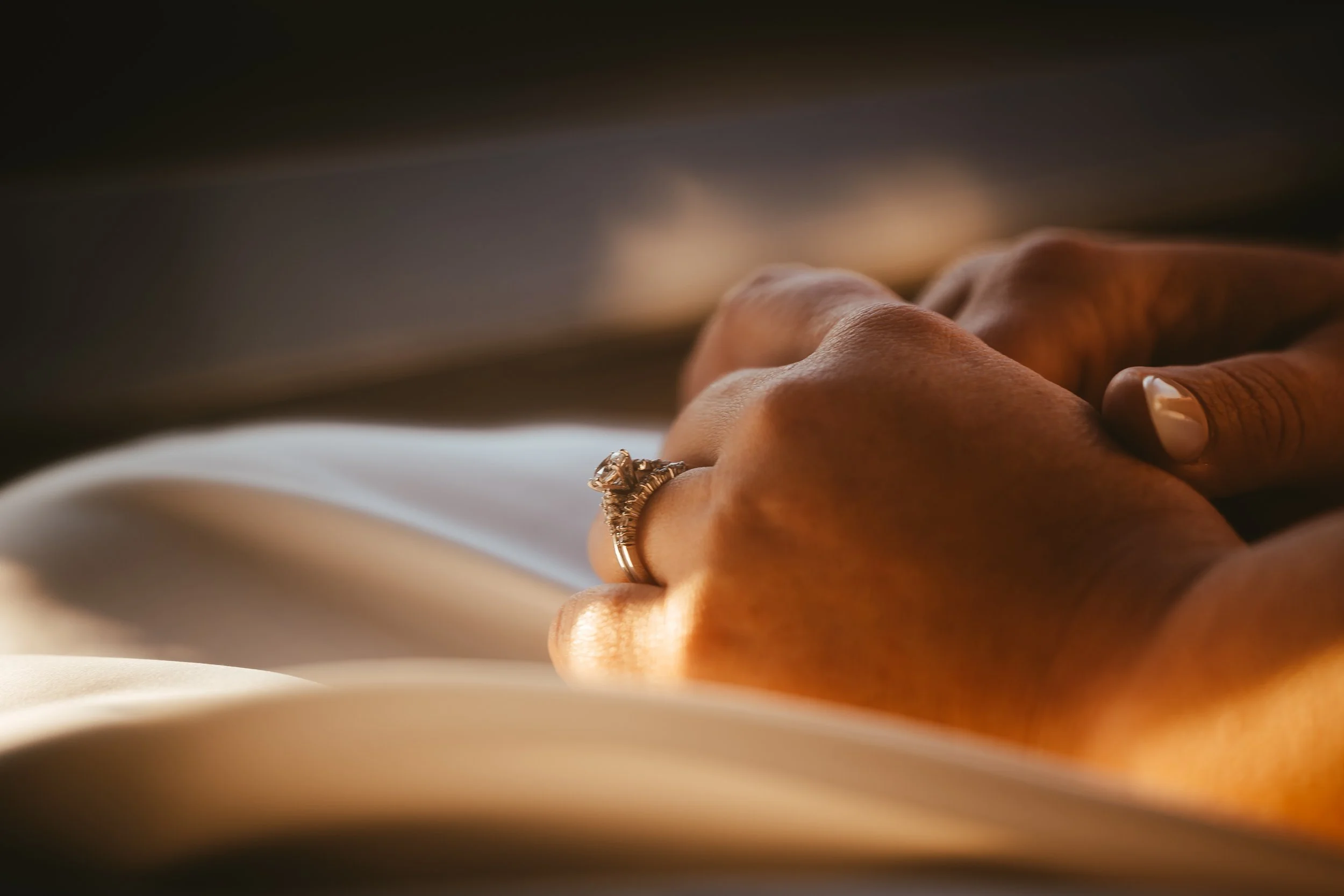 Close-up of a hand wearing a large gemstone ring, resting on an open book, with a dark background.
