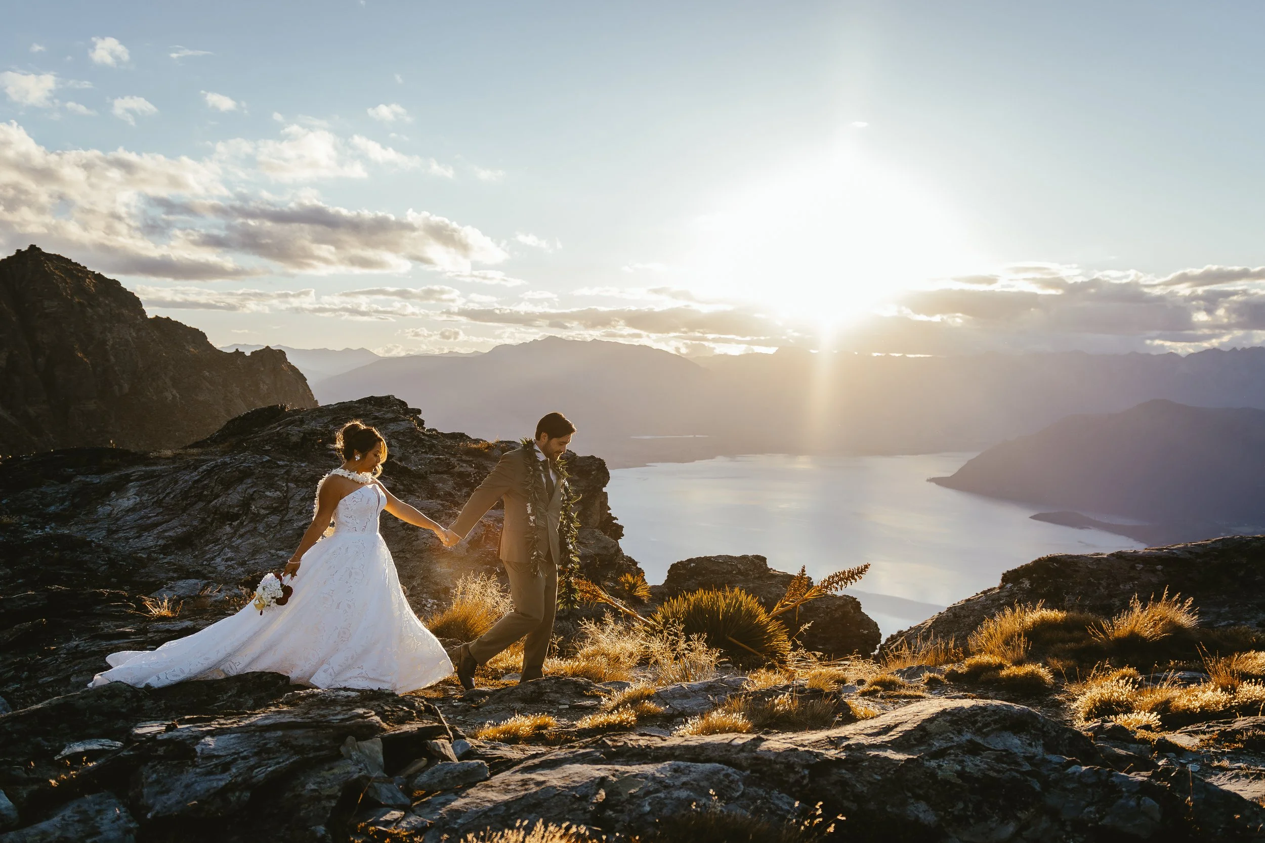 A bride and groom walking hand in hand on a rocky terrain during sunset with mountains and a lake in the background.