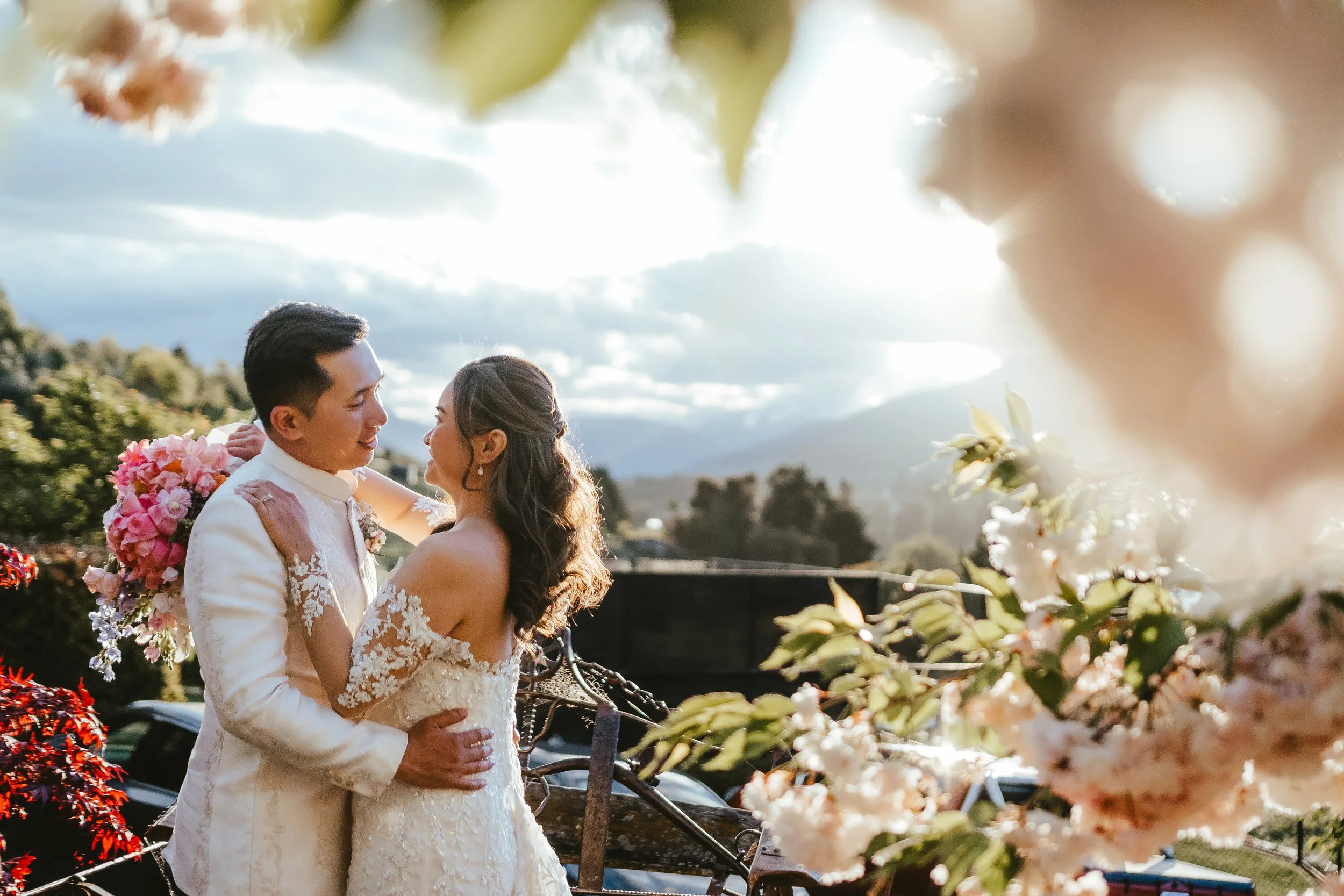 A newlywed couple stands outdoors, gazing into each other's eyes. The groom is wearing a white suit with lace details, and the bride is in a white lace wedding dress. They are surrounded by flowers and a scenic backdrop of hills and a cloudy sky.